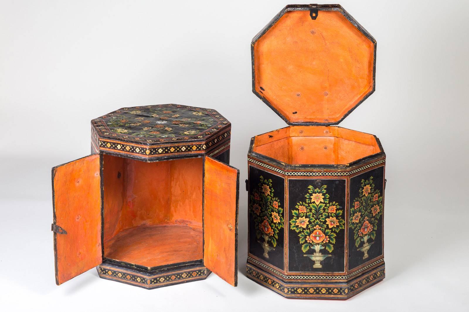 Pair of Anglo Indian octagonal colorful papier mâché painted low end tables decorated with urns containing flowering plants on a black background. One has a hinged top that lifts to reveal an orange interior; the other has hinged double doors on the