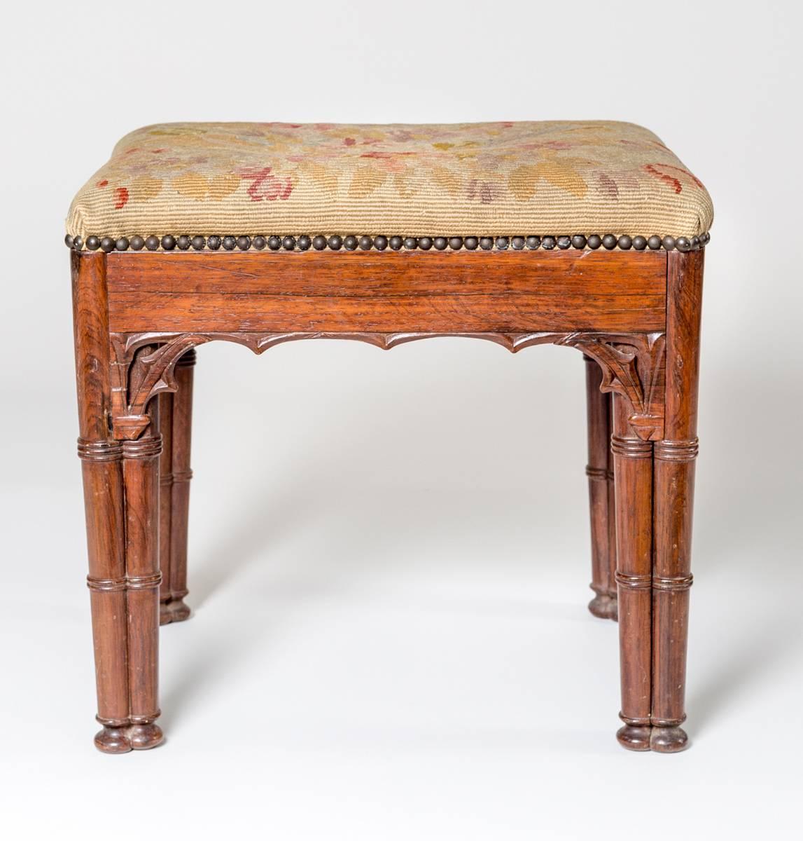 Victorian rosewood Gothic Revival stool, the apron with arched moldings, supported by tri-cluster column legs with ring turnings, the seat covered in a needlework decorated with two birds and flowers with brass nailheads.
