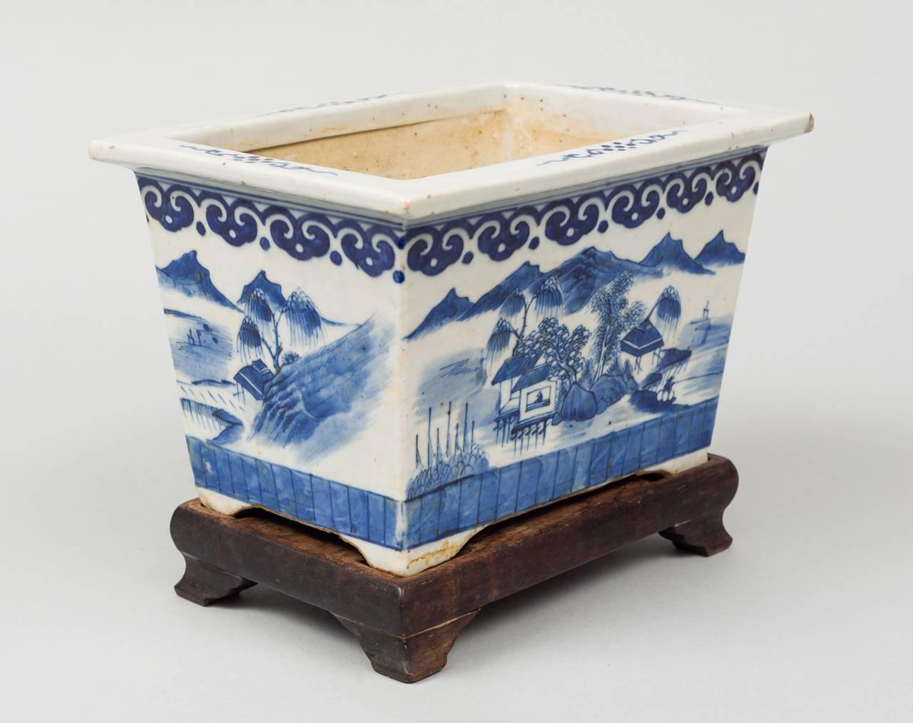 Chinese export porcelain rectangular blue and white jardiniere on wooden stand. It is decorated at the top with a ruyi head border and the body with exotic houses, mountains, trees and figures and set on small bracket feet.