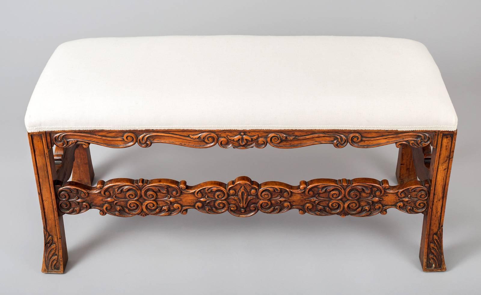 French walnut bench with shaped aprons and stretchers on all sides.  The aprons and stretchers are boldly carved with C-scrolls and double C-scrolls.  The squared legs are splayed with carving at the bottom of the feet.  Upholstered in an off-white