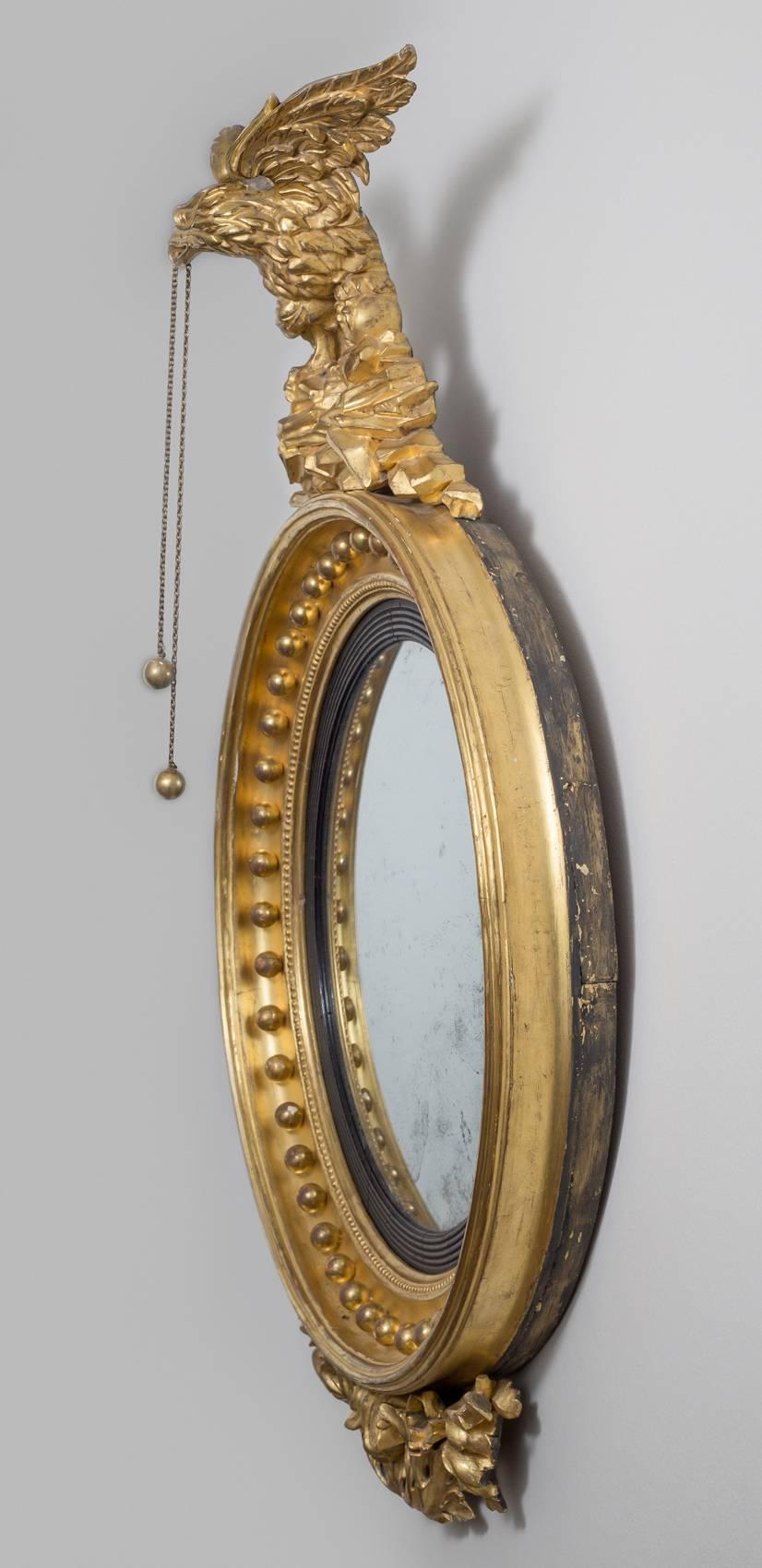 Regency giltwood convex wall mirror, the circular original mercury plate and ebonized reeded slip set within a molded concave frame with ball spacers, surmounted by a spread winged eagle with balls and chains hanging from its beak, perched on a