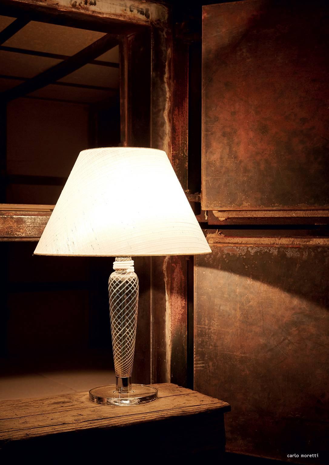 Table lamp with Murano glass column, grey/white spiral transparent glass base, a truncated cone-shaped fabric shade, and metal fixtures in polished chrome. The lamp was designed by Carlo Moretti in 1995.


Carlo Moretti: An artisan