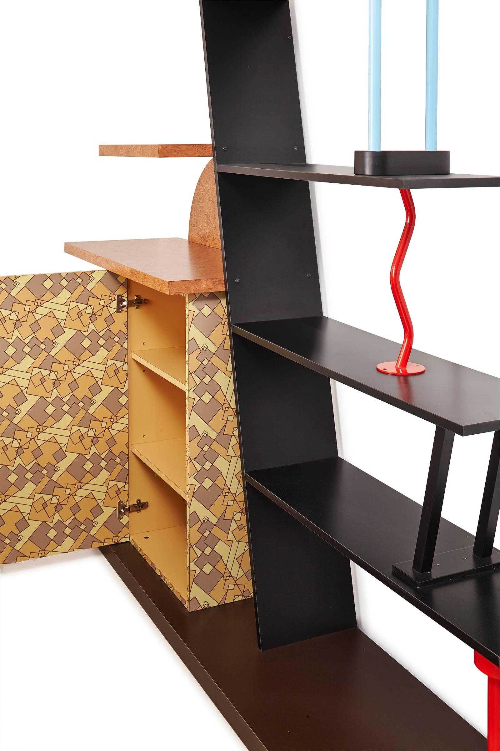 The piece consists of six shelves with a one-door cupboard decorated in plastic laminate with a geometric pattern. Its form is made up of both straight and curved lines punctuated by diagonal and zigzag structural elements. 

Malabar is a perfect