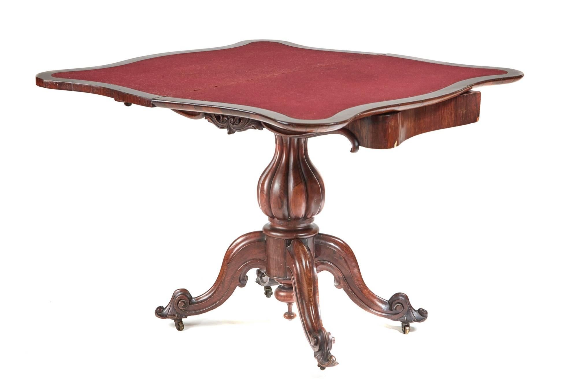 A good quality Victorian rosewood serpentine shaped fold over card table with swivel top, carving around the frieze, central reeded baluster shaped column standing on four sweeping carved cabriole legs with sea scroll on the ends, red baize