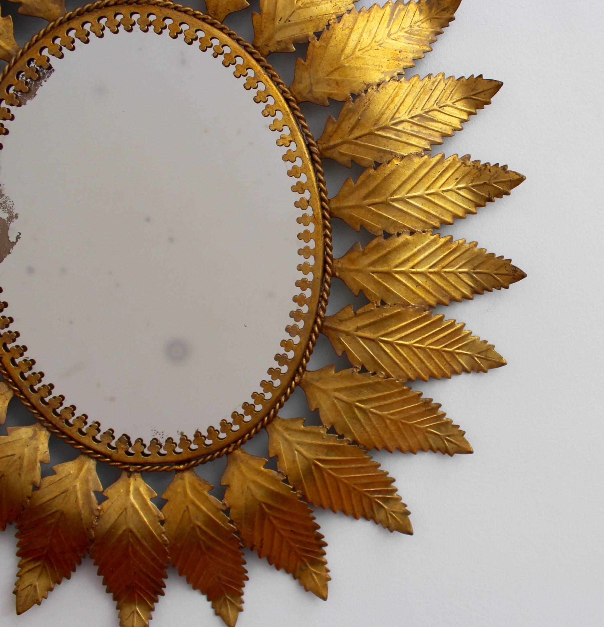 Spanish gilt metal sunburst mirror (circa 1950s) with leaf motif rays and cross-pattern mirror border. In fair vintage condition commensurate with age and use. Some authentic age spots, blemishes and degradation appear on mirror surface reflecting