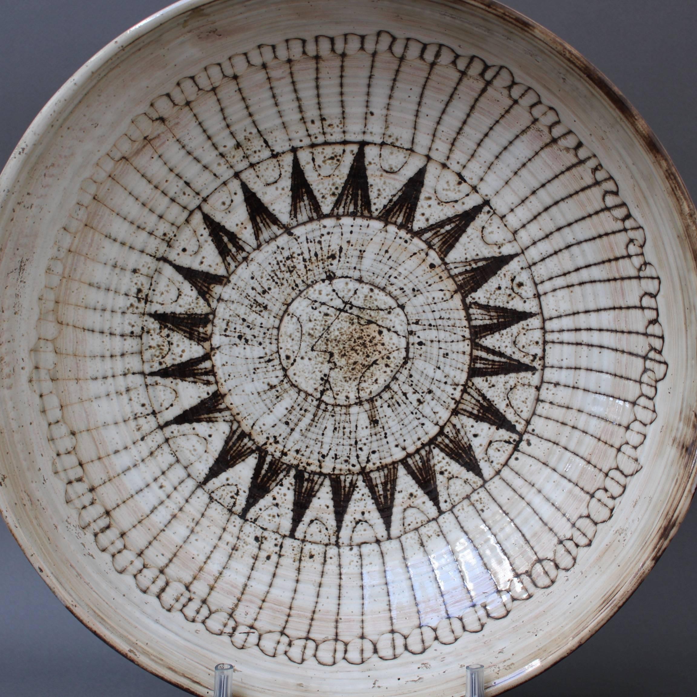 Large midcentury decorative bowl (circa 1960s) by Jacques Pouchain - Atelier Dieulefit. This decorative bowl has as its centre, a large sunburst motif in brown over a wider glaze of dusty white. There is a stylised face in the middle of the sun.