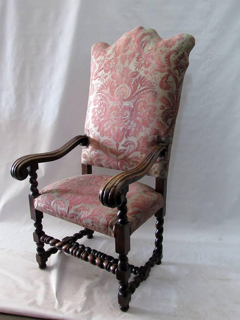 A tall carved wood Louis XIII style armchair upholstered in Fortuny. The beautifully shaped crest rail above an upholstered back with crowned seat below flanked by ribbed carved arm rails that rest on barley shaped posts that display the same style