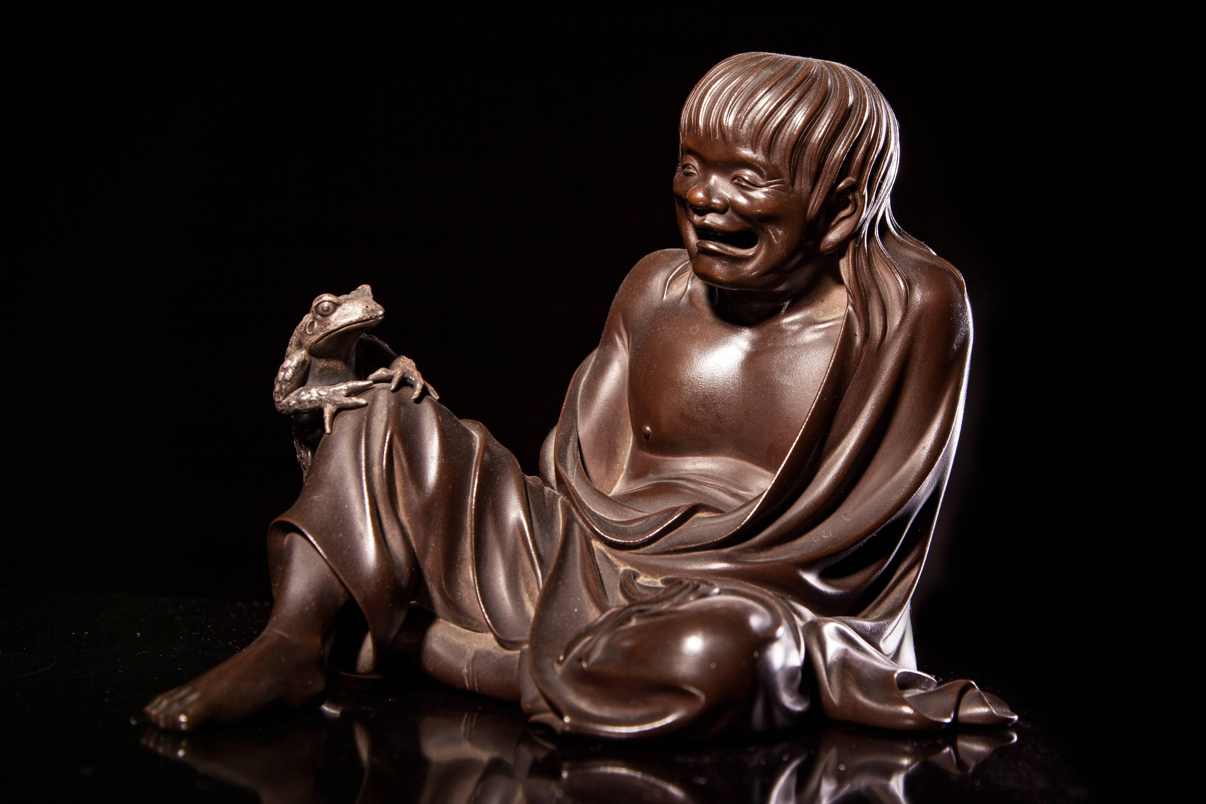 A Japanese late Meiji Taisho period bronze of Gamma Senin as a seated loose robed man, a separate white metal toad on his bent and raised right knee,
on lacquered wooden plinth,
signed Kano Seiun to base,
Measures: H 15.5 cm W 21.5 cm.