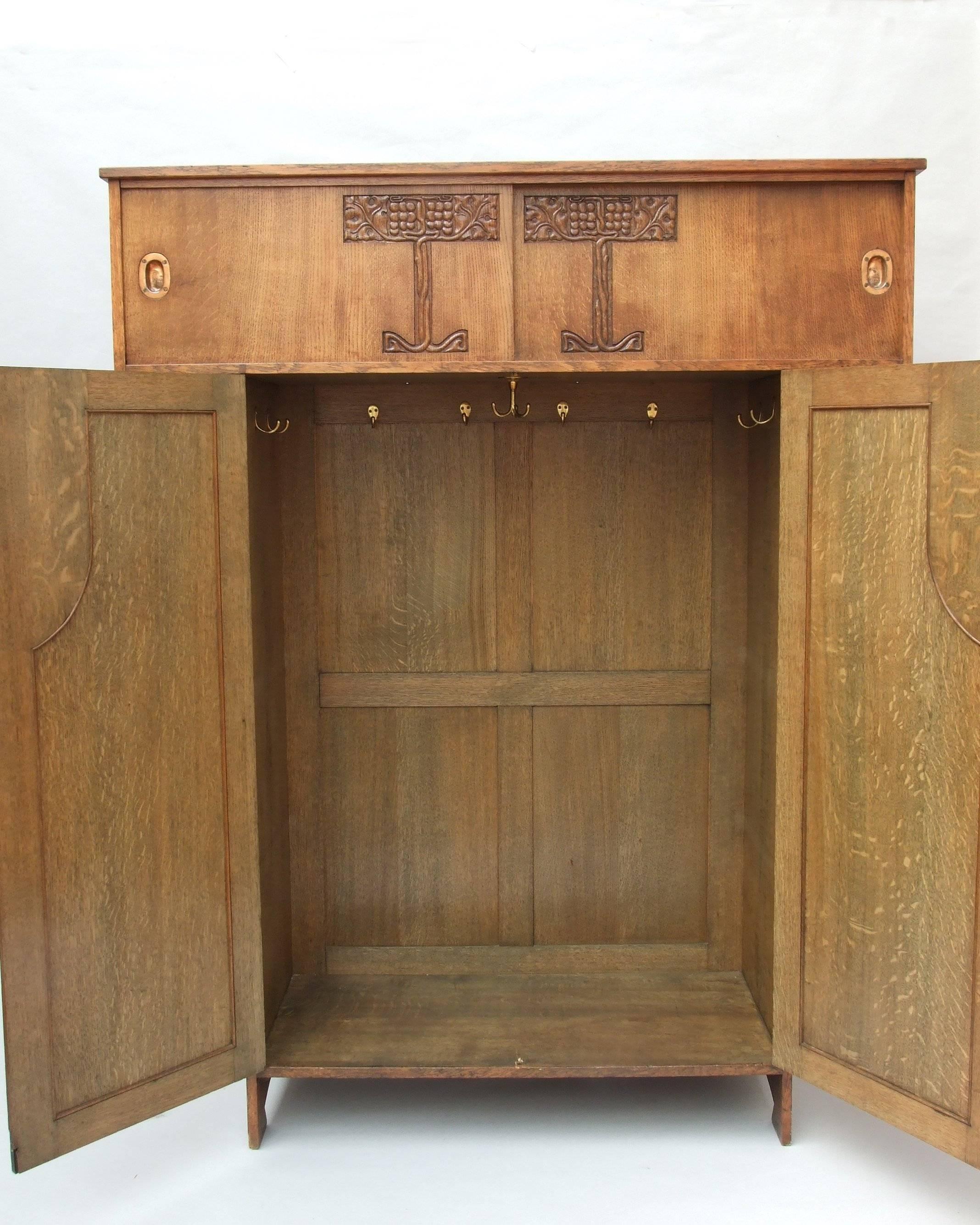 A Liberty oak Kimono-shaped wardrobe, overhanging top cupboard with two sliding doors, inset copper handles, stylized leaf and berry carved decoration to each door, over two shaped panelled doors with fine bronzed patinated latch on roundel with