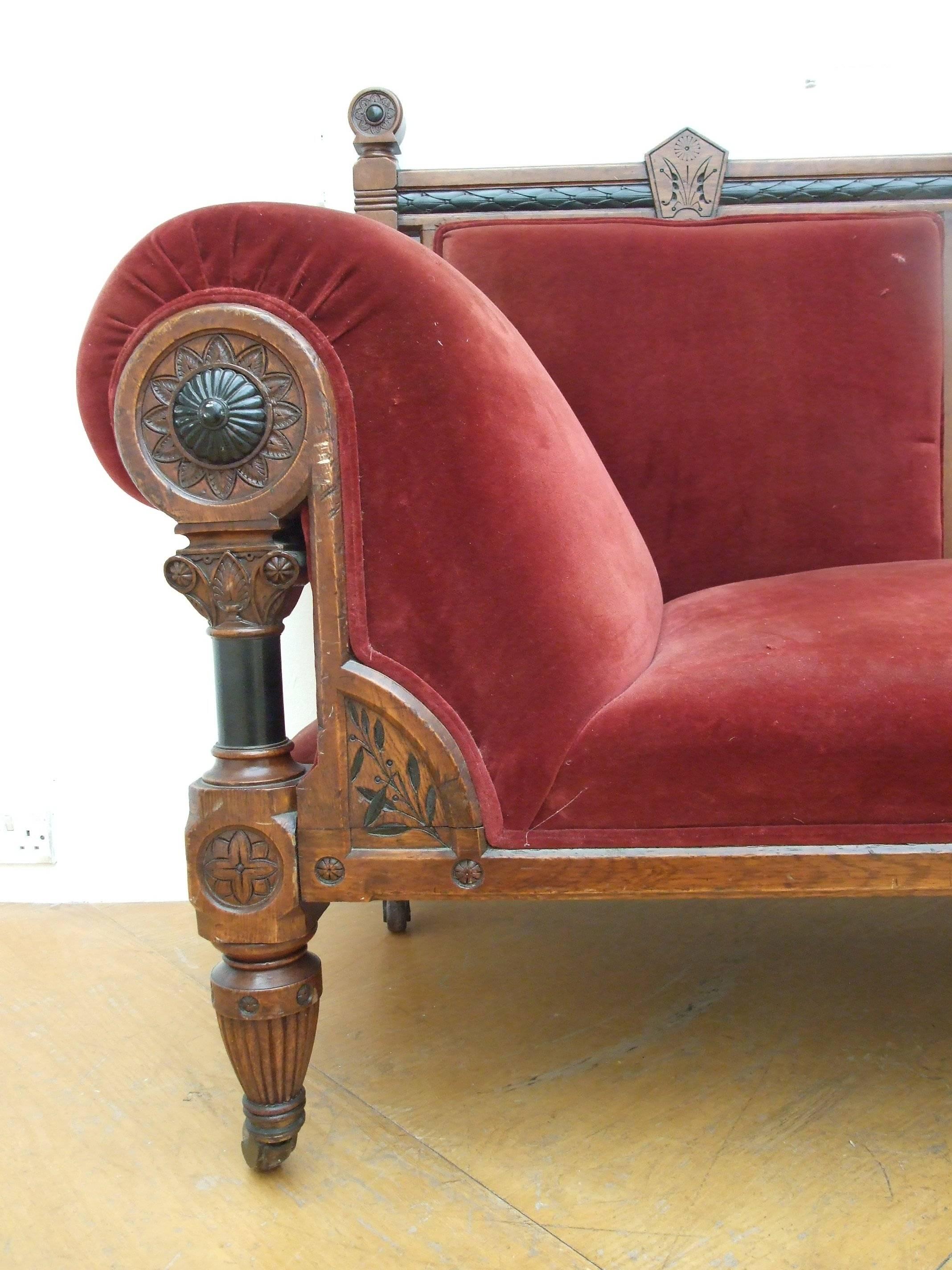 A pollard oak Aesthetic Movement chaise longue, with spindles and carved frieze in ebony, early Classical finials to the uprights with carved stylized flower decorations, reeded tapering legs on brass and porcelain casters, upholstered in burgundy