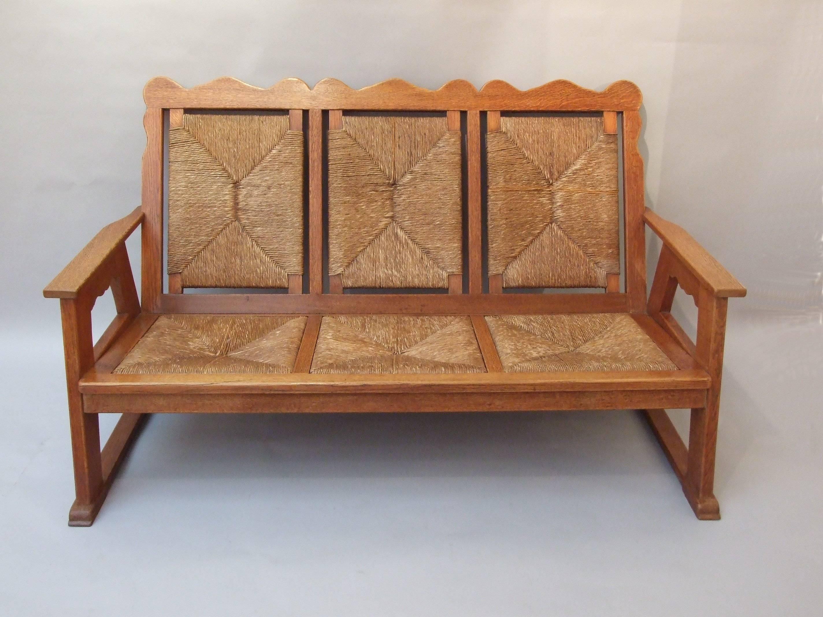 A rare Arts & Crafts oak three piece suite, two arm chairs and a three-seater settle, with shaped backs, sides and apron, stepped sledge feet, original rush backs and seats, by William Birch of High Wycombe, design attributed to E G Punnett,