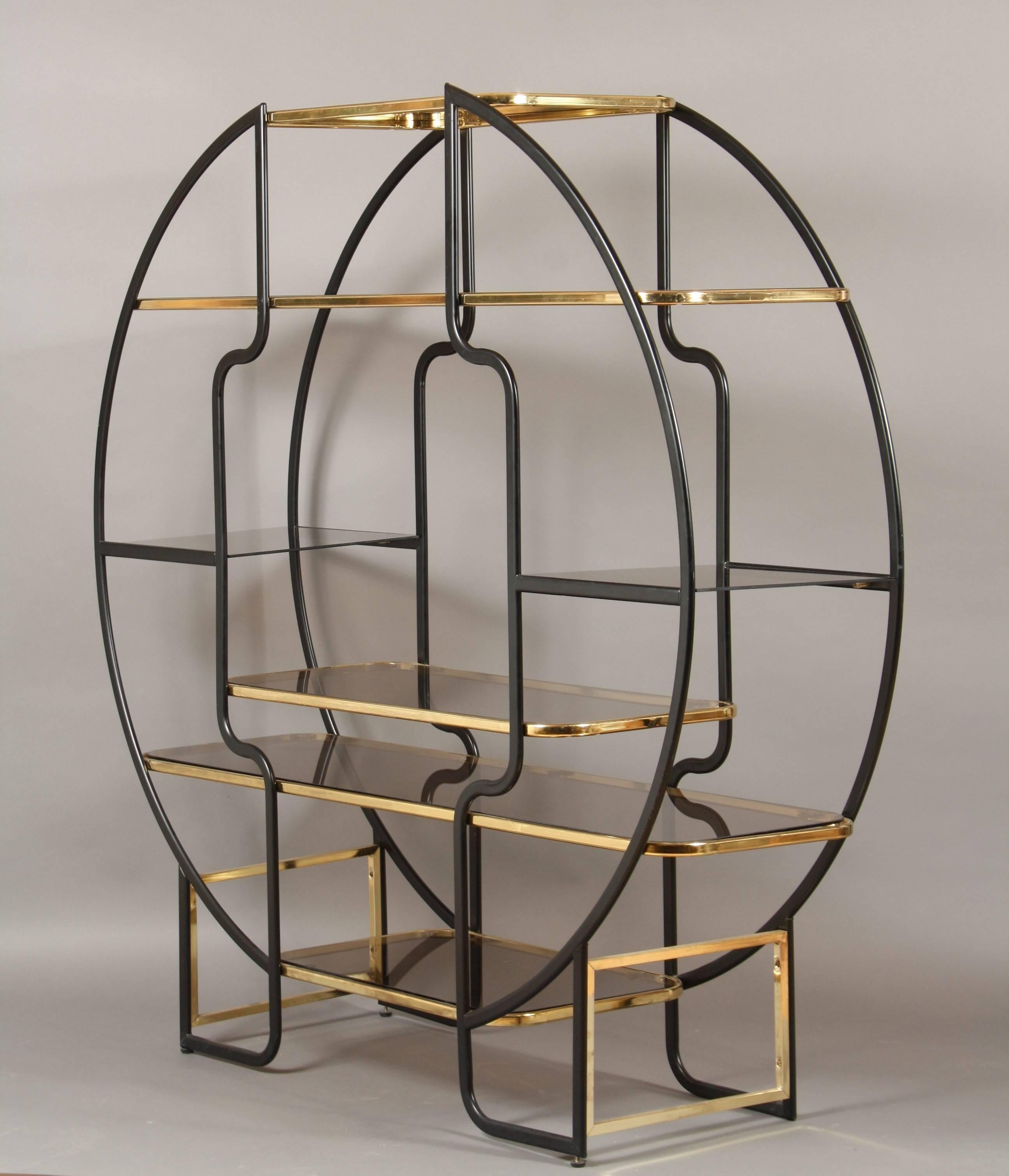 Art Deco bookcase from the 1970s.
Its two-sided frame constitutes a circle and is made of black colored metal. The two half-circles are tied together by shelves that are made of smoke-colored glass with a brass frame.
This extraordinary piece