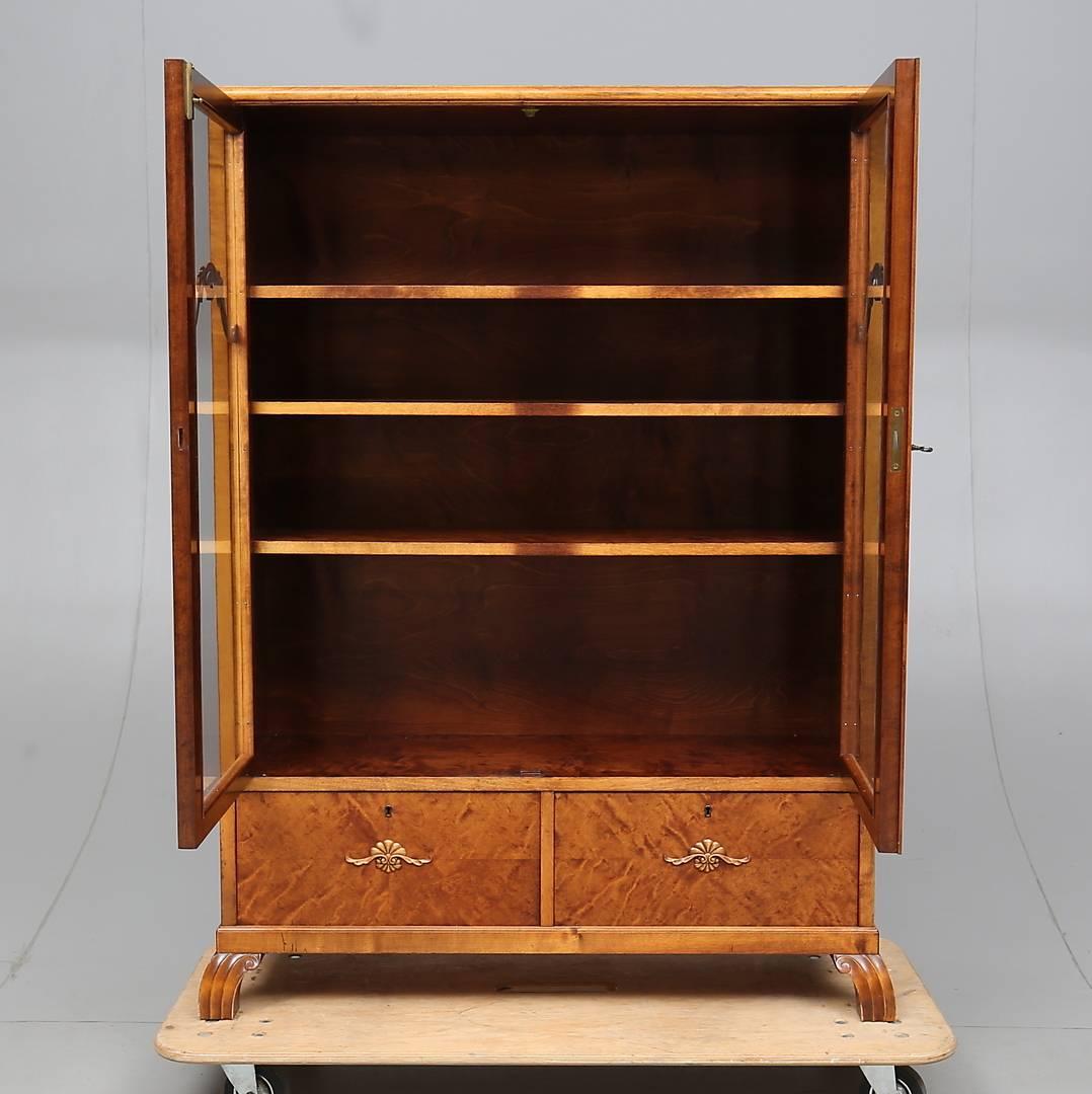 Beautiful Swedish antique Art Deco cabinet in quilted gold birch veneers with carved detailed on the glazed doors, deco feet and inlaid marquetry on the lower doors, circa 1920s

Three shelves inside the cabinet and the bottom doors unusually open