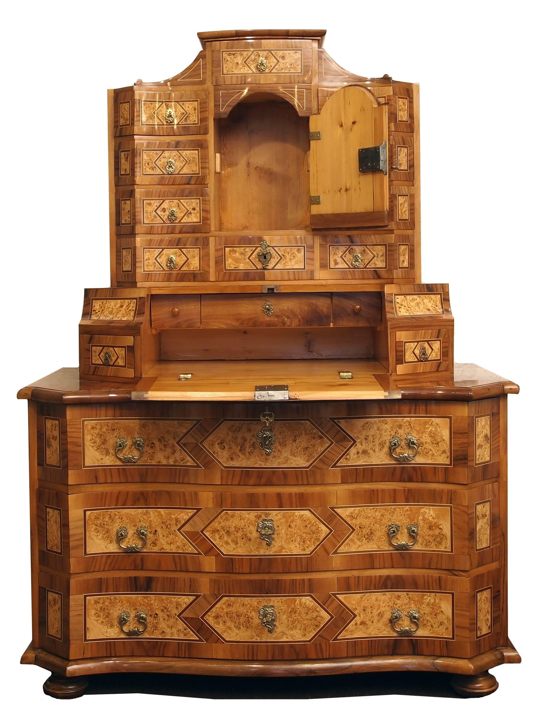 The tabernacle secretary desk stands on low round feet and is richly decorated with noble wood inlays. The chest of drawers bottom part is concave curved and has three drawers. The writing surface is at a height of 88 cm. Above is a high top part