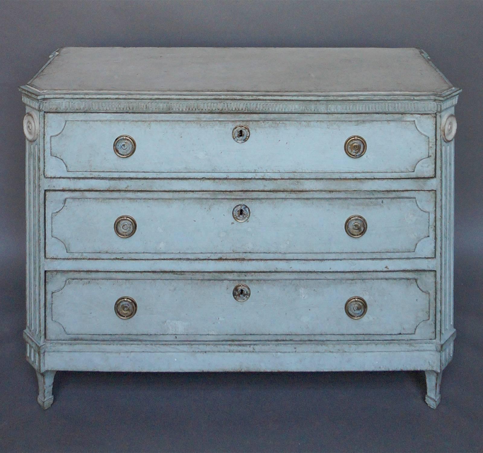 Chest of three-drawers, Sweden, circa 1840, in pale blue paint and with a reeded panel under the shaped top, canted corner posts with fluting, and raised panels on each drawer front. Brass pulls and escutcheons.