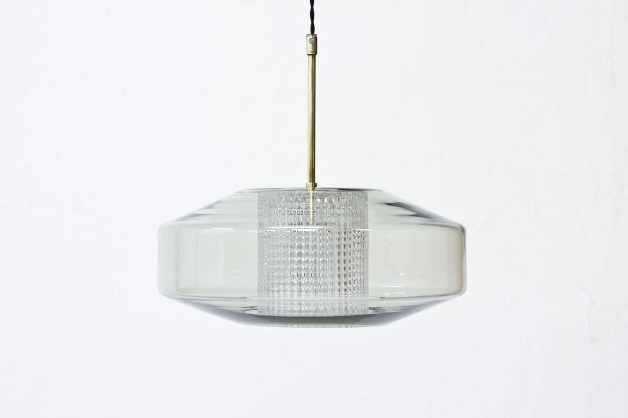 Pendant lamp designed by Carl Fagerlund for Swedish glass maker Orrefors during the 1960s.
Flattened sphere shape in clear grey tinted glass with a cylinder internal diffuser in clear pressed glass. Brass fixture. Braided black textile cable. Very