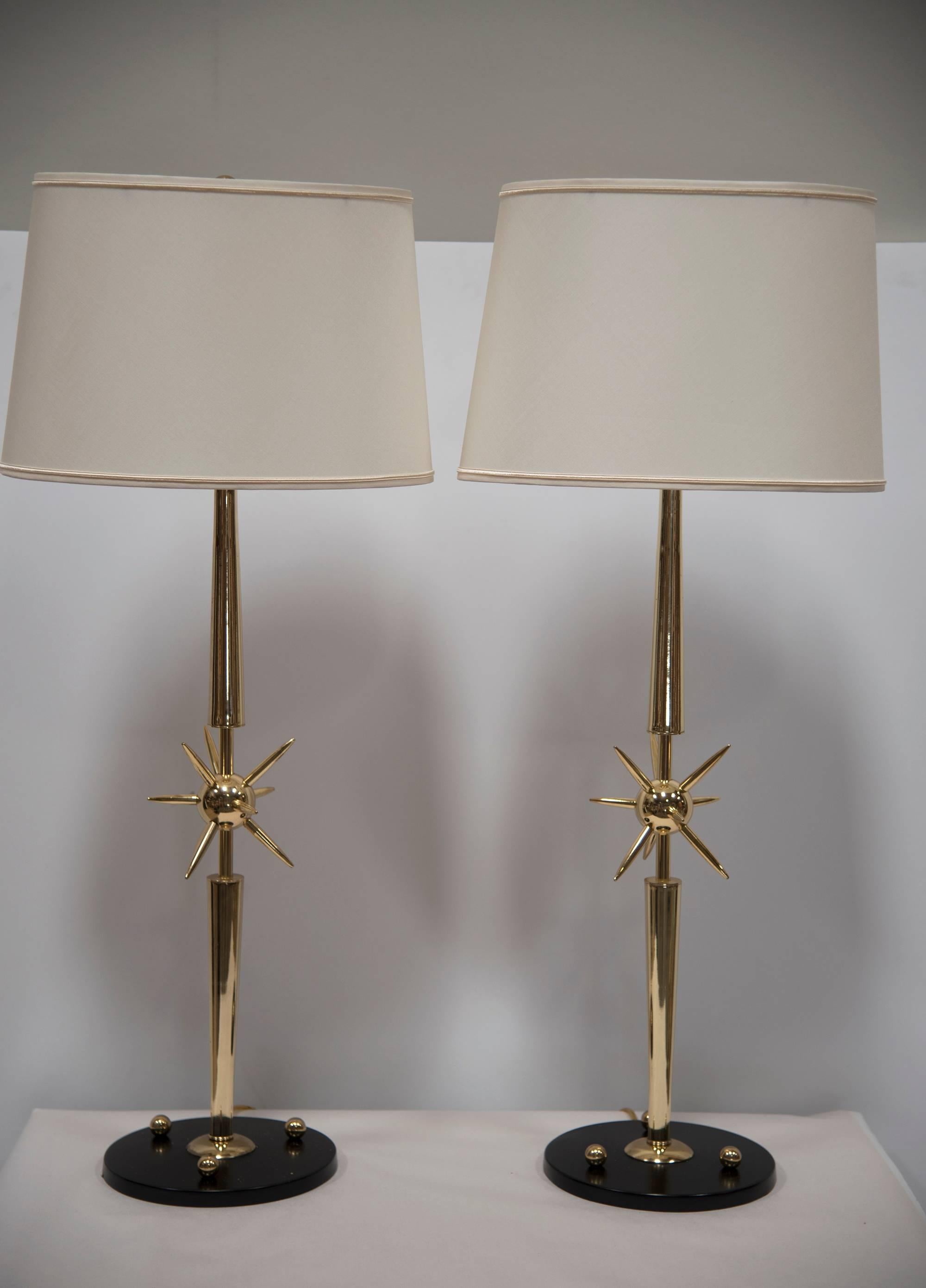 A pair of tall, elegant, conversation piece brass table lamps in the form of starbursts in the centre of each column. Brass with lacquer black base accessorised with studs. Lamp shades are pristine condition with a high-end linen finish and gilt