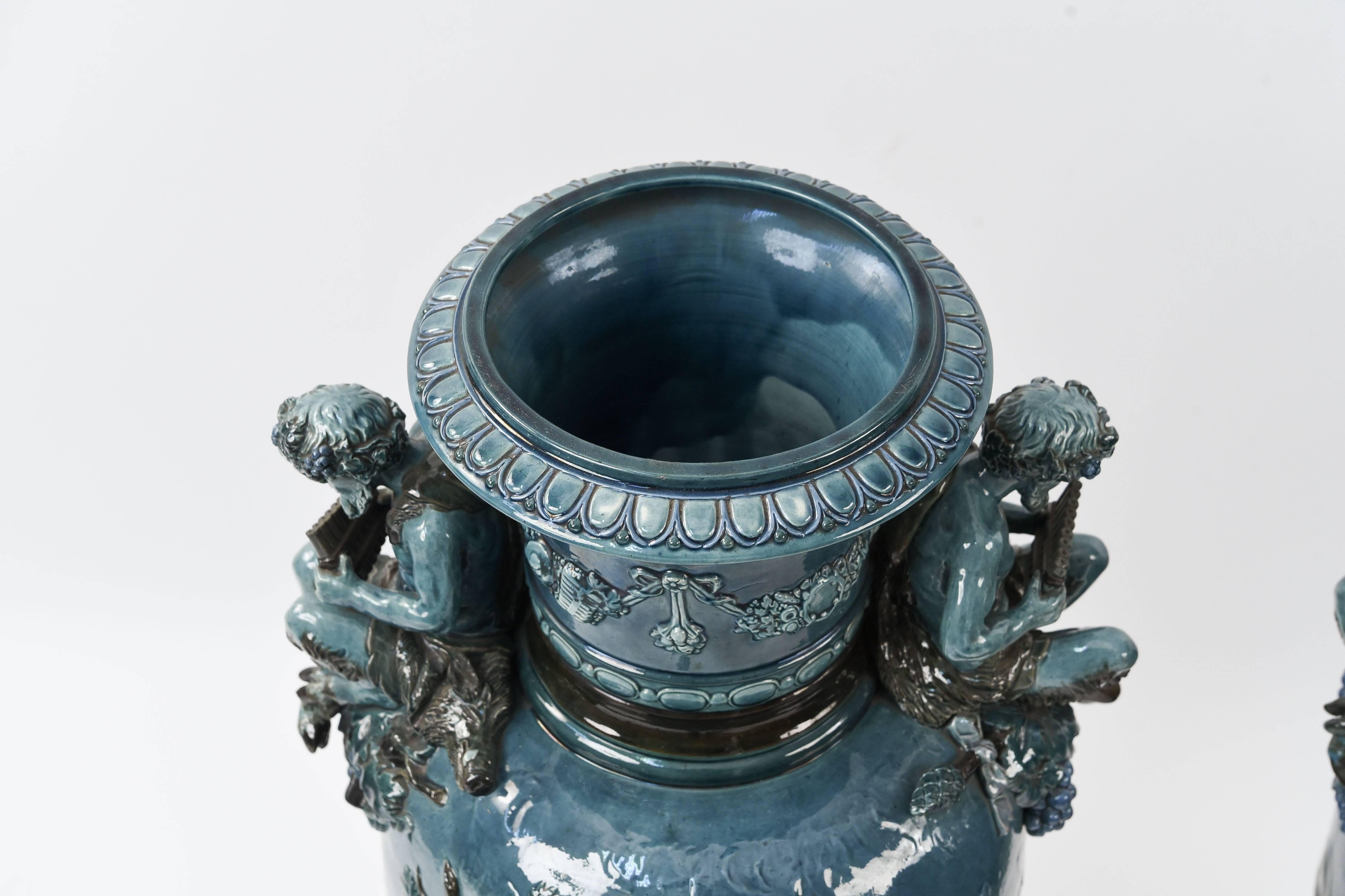 A wonderful, large pair of teal French majolica urns. Featuring two applied pan figures on each side of the openings, and a mother and child scene in relief on the bodies of the urns. The color and size make for a very striking pair.