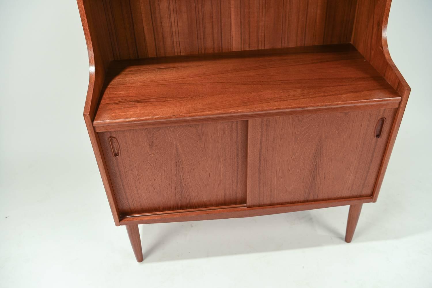 This Danish midcentury bookcase in the manner of Johannes Sorth for Nexø is made of handsome teak wood and features multiple shelves and a compartment below with wonderful recessed handles for storage. Great display piece.