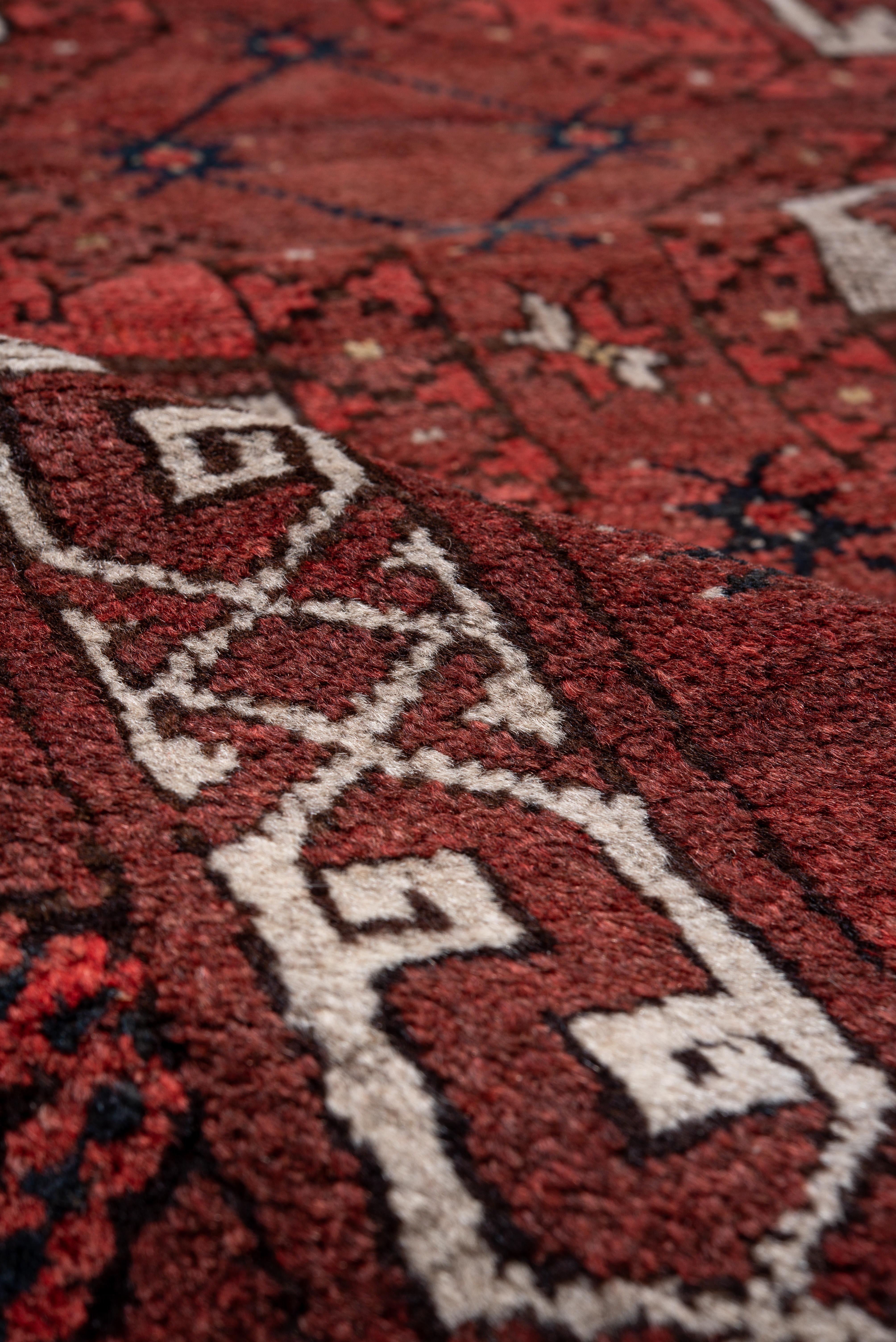 This vintage Afghan rug presents a rich crimson field punctuated by rows of traditional gul motifs in earthy brown, ivory, and charcoal hues. Each gul features quadrant-style symmetry, echoing the tribal heritage of the region, while subtle flecks