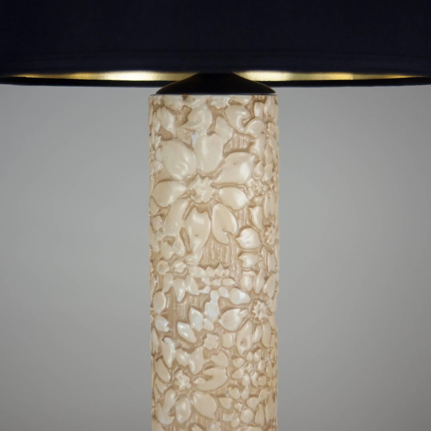 Cylindrical ceramic table lamp embossed with floral design and with black metal base. Lamp has been professionally rewired with black silk cord and has a new black linen opaque shade with gold interior. Shade measures 17