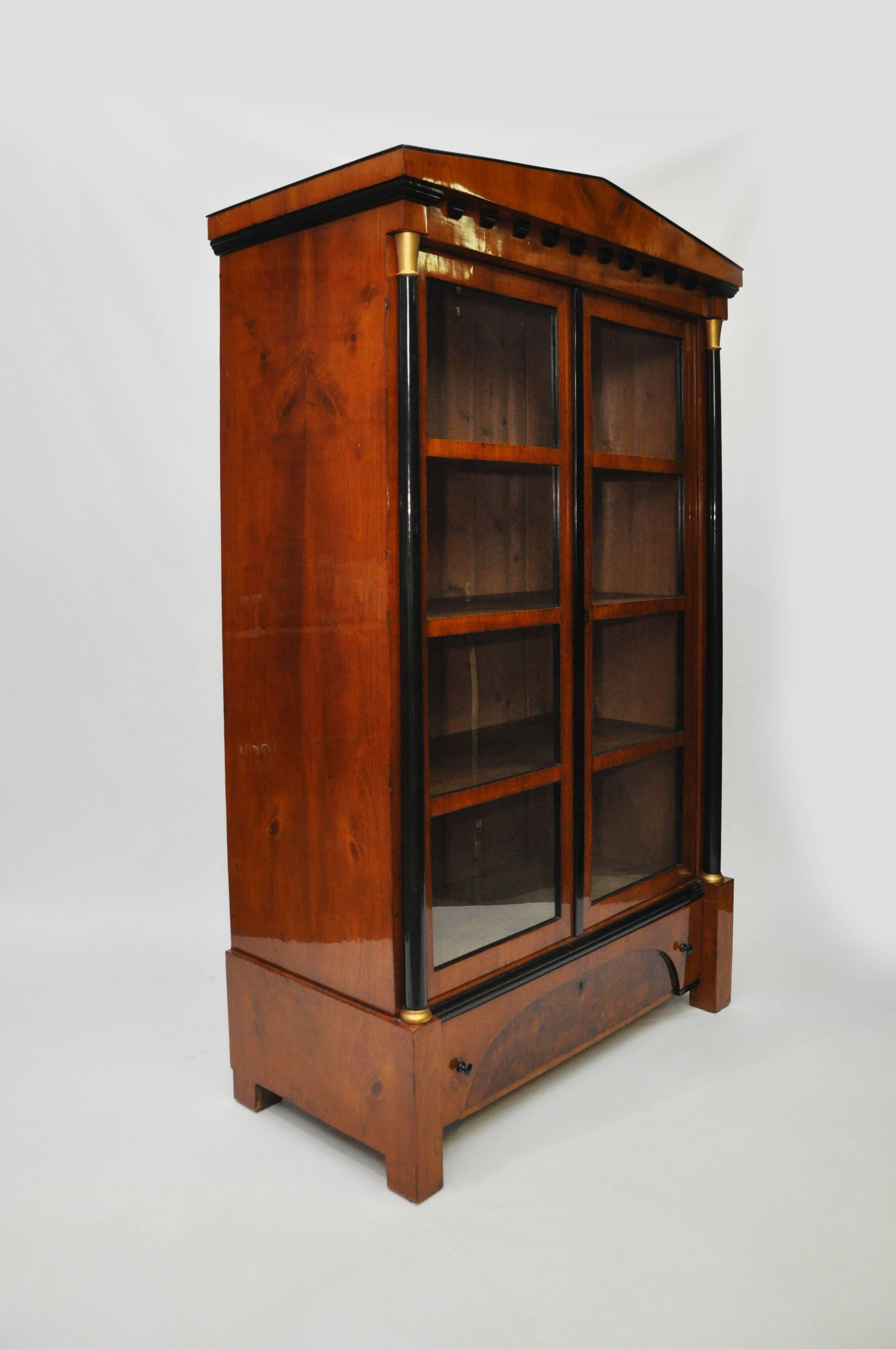 Stunning Biedermeier bookcase with dramatic architectural style pedimented top. Warm cherry veneer on pine with ebonized column and korbels. Matte gold painted capitals. Burl walnut veneer on the front of the drawer. Stationary pine shelves. The