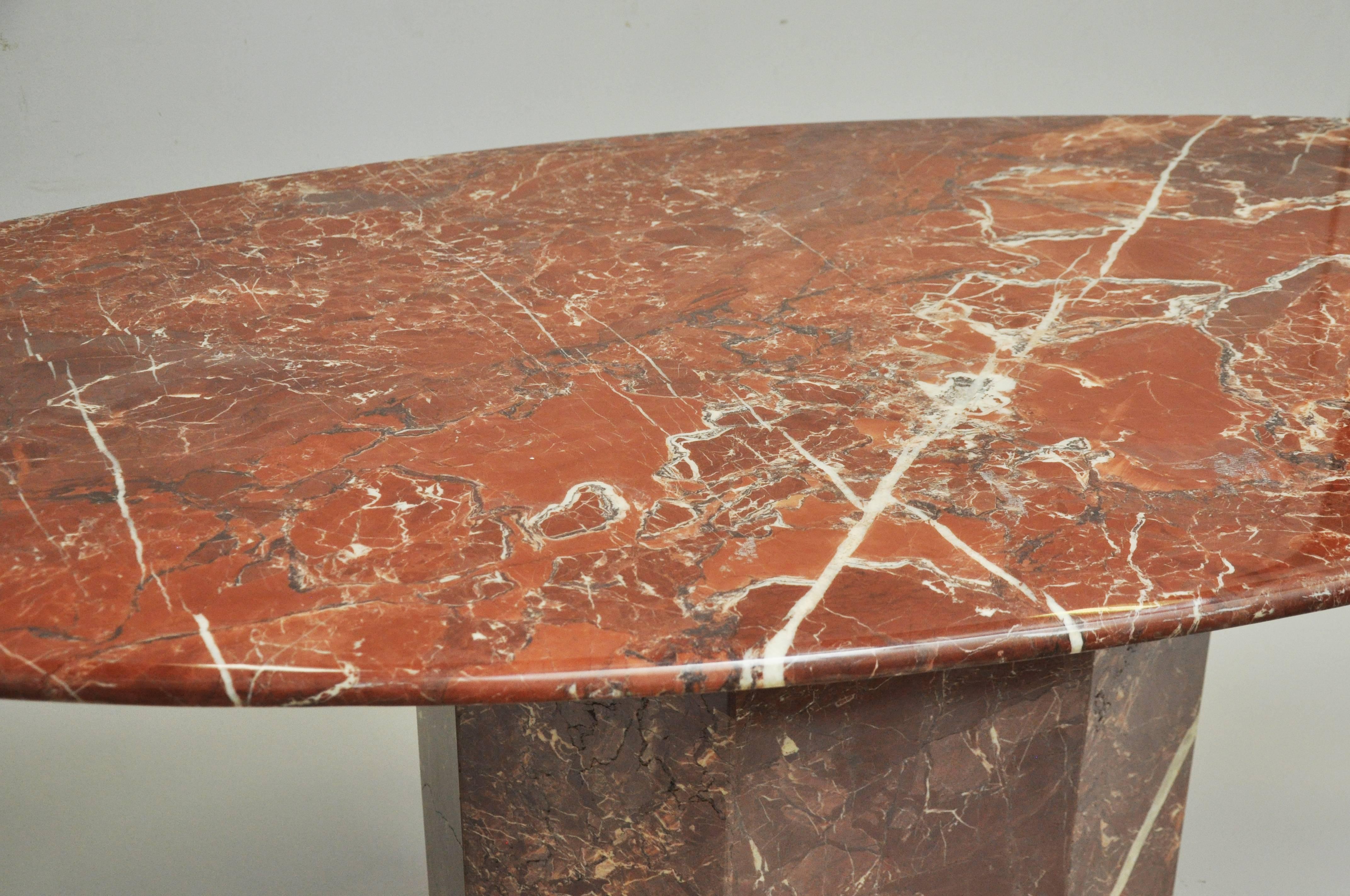 Midcentury highly figured marble top oval dining table resting on octagonal matching marble base. Table is in red tones with striations and veining of off-white and grays.