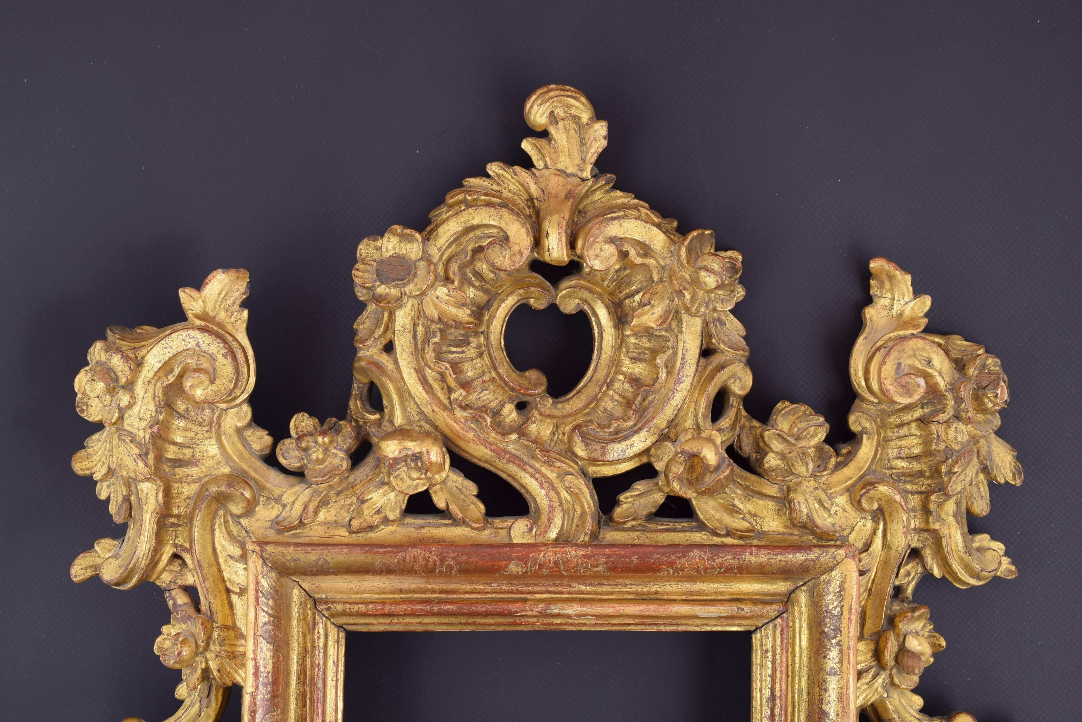 Rococo carved wood frame, 18th century.
Carved wood rectangular frame decorated with a symmetrical composition of “rocaille”, architectural elements and leaves, drawing curves, as is usual in Rococo art, with some small flowers. 
Nowadays, the