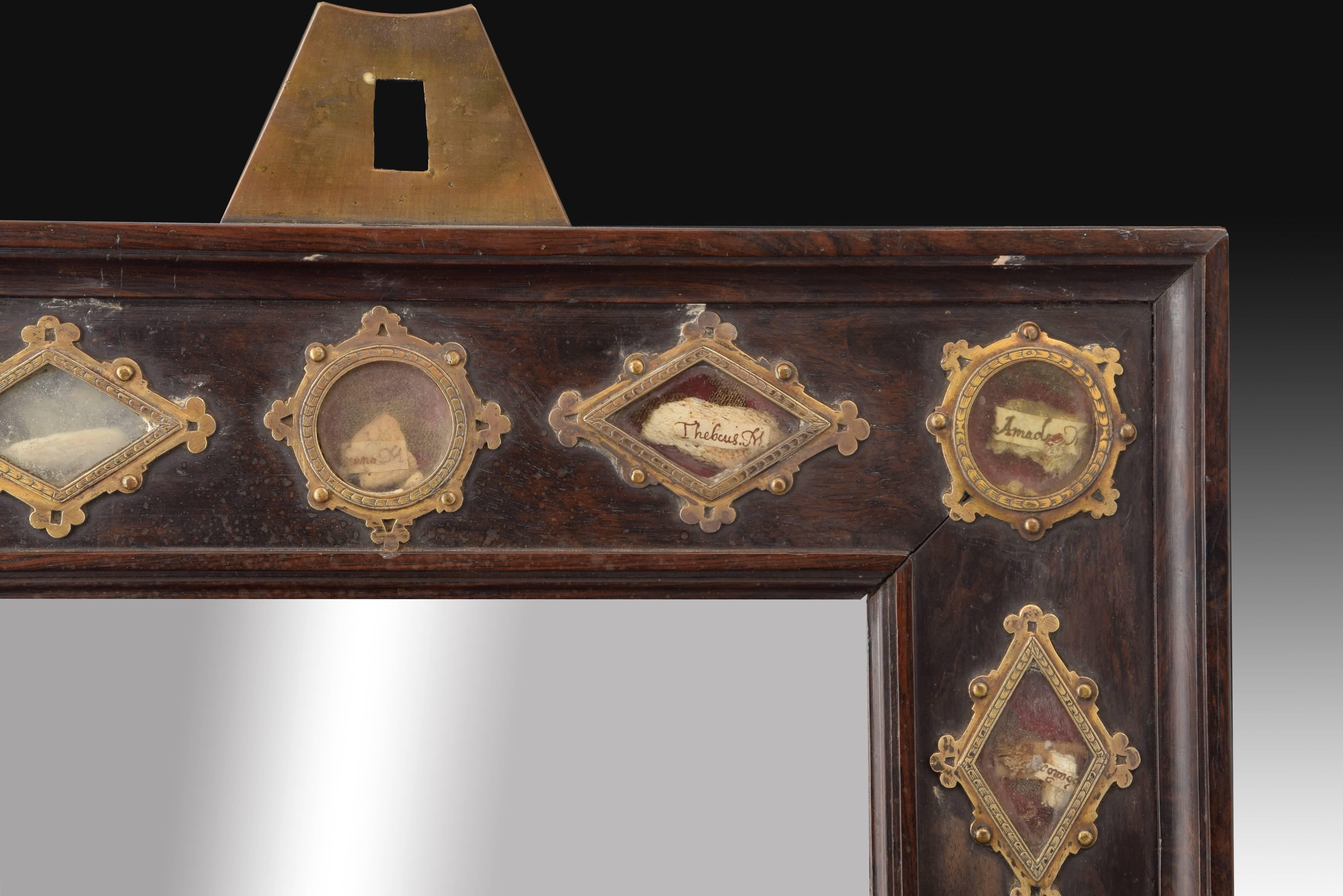 Rectangular frame of carved ebony wood decorated with fine moldings on the edges of it. It has a metal piece on the top to place it on the wall and, in the central part of it, a series of spaces reserved for different relics. These present several