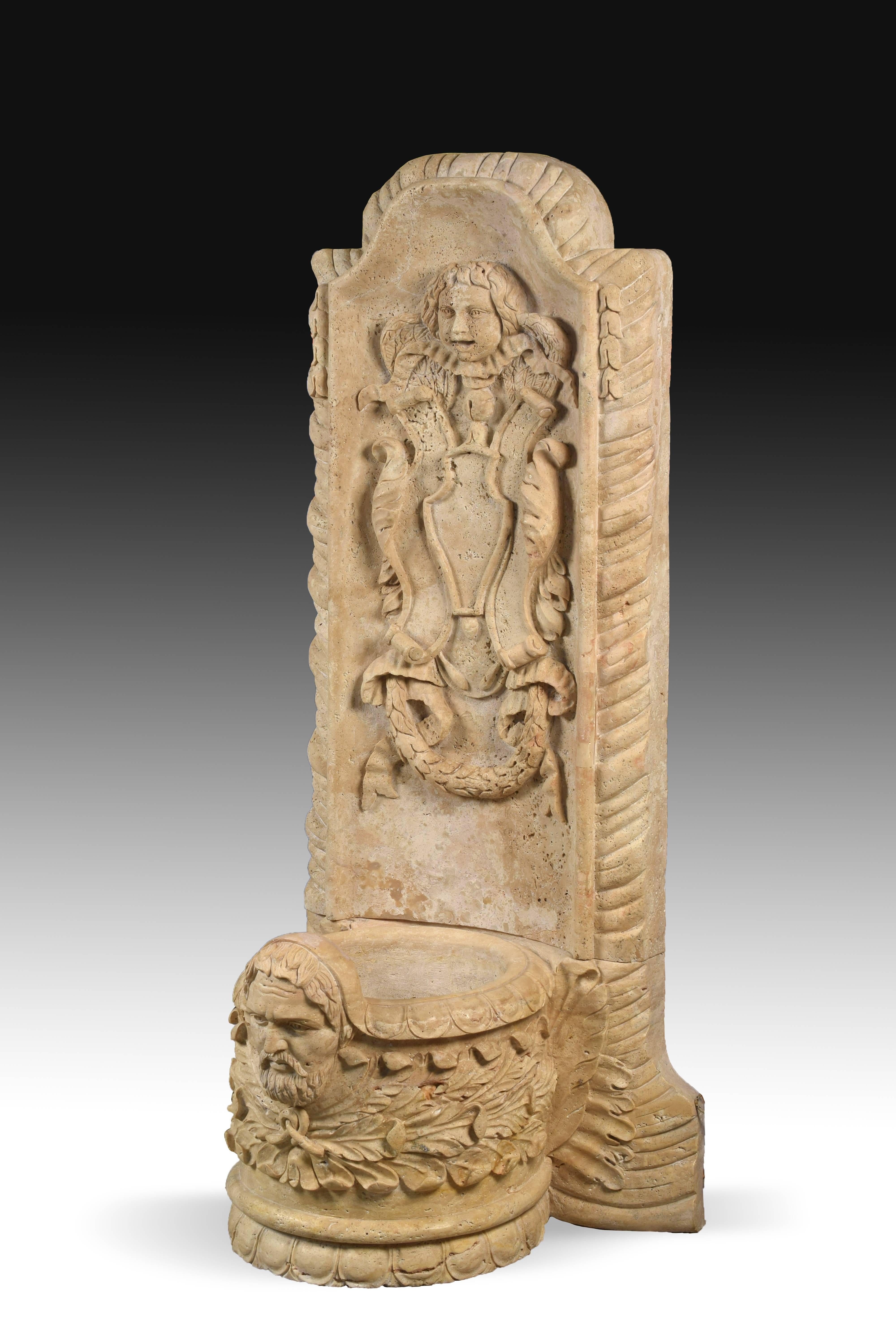 Fountain made in carved travertine with low cup, decorated with a bust and vegetal elements and a high panel to fix it to the wall, also decorated with high relief carvings of classicist influence. Although the elements show the influence of models