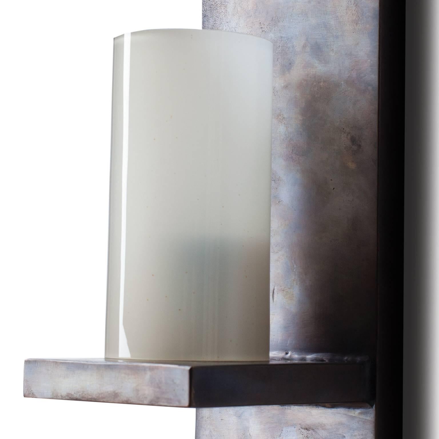 The Roca sconce is made of cast bronze and has a hand applied patina that gives a rich warm depth to the metal. The light source is housed in an opaque glass cylinder. Cast bronze has natural markings that are inherent to the process and  techniques