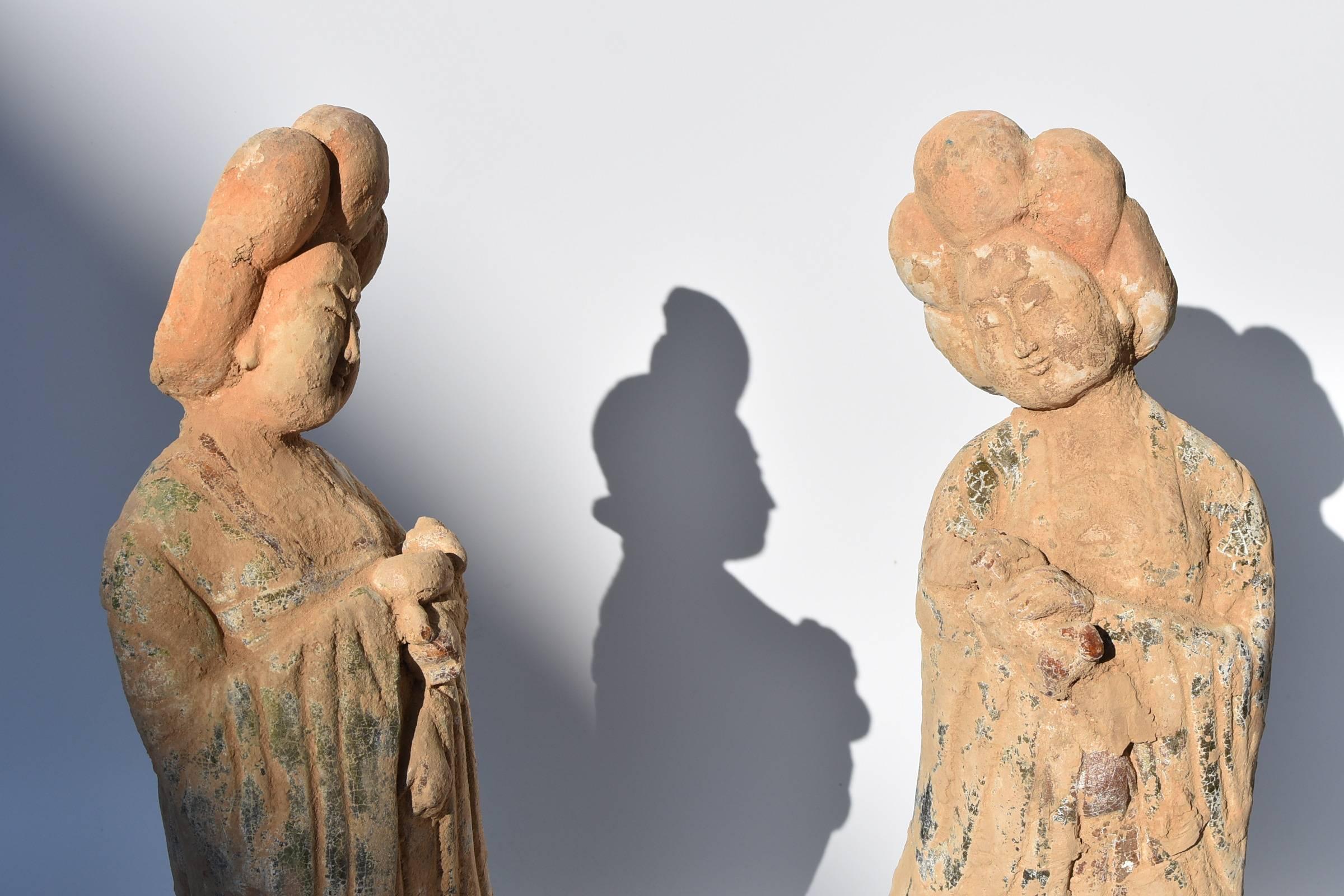 A pair of terracotta court ladies in the style of Tang dynasty. The ladies have the traditional Tang full faces and beautiful features. Their body postures are demure and graceful. They are both carrying a puppy, with another puppy at each of their