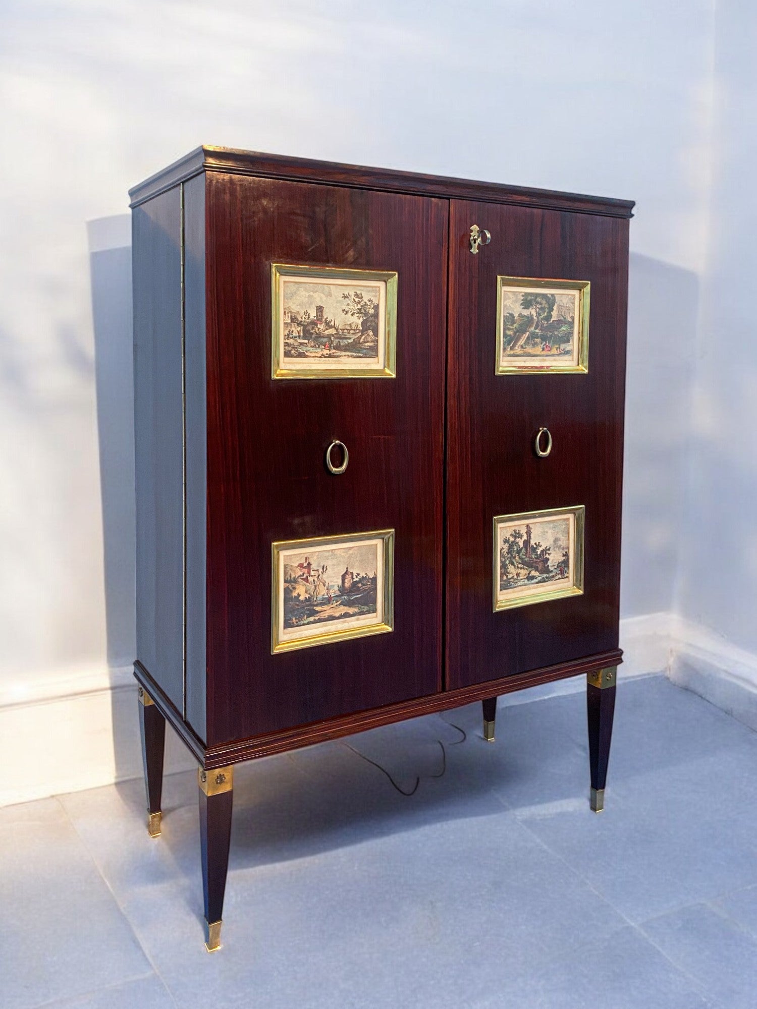 Este elegante mueble bar, diseñado por Paolo Buffa en la década de 1950, es un verdadero ejemplo del diseño italiano de mediados de siglo. La parte frontal presenta cuatro paneles de madera dorada, enriquecidos con grabados del siglo XVIII que