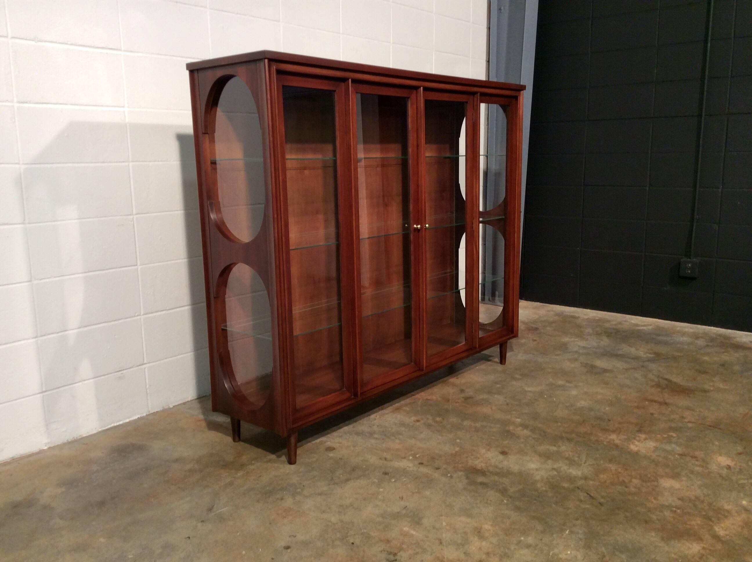 Mid-Century Modern walnut and glass display cabinet by Kent Coffey on added pencil legs. Very unique with see through side panels. Perfect for displaying china, pottery, or even purses and shoes. The cabinet does have a light in the top so you can