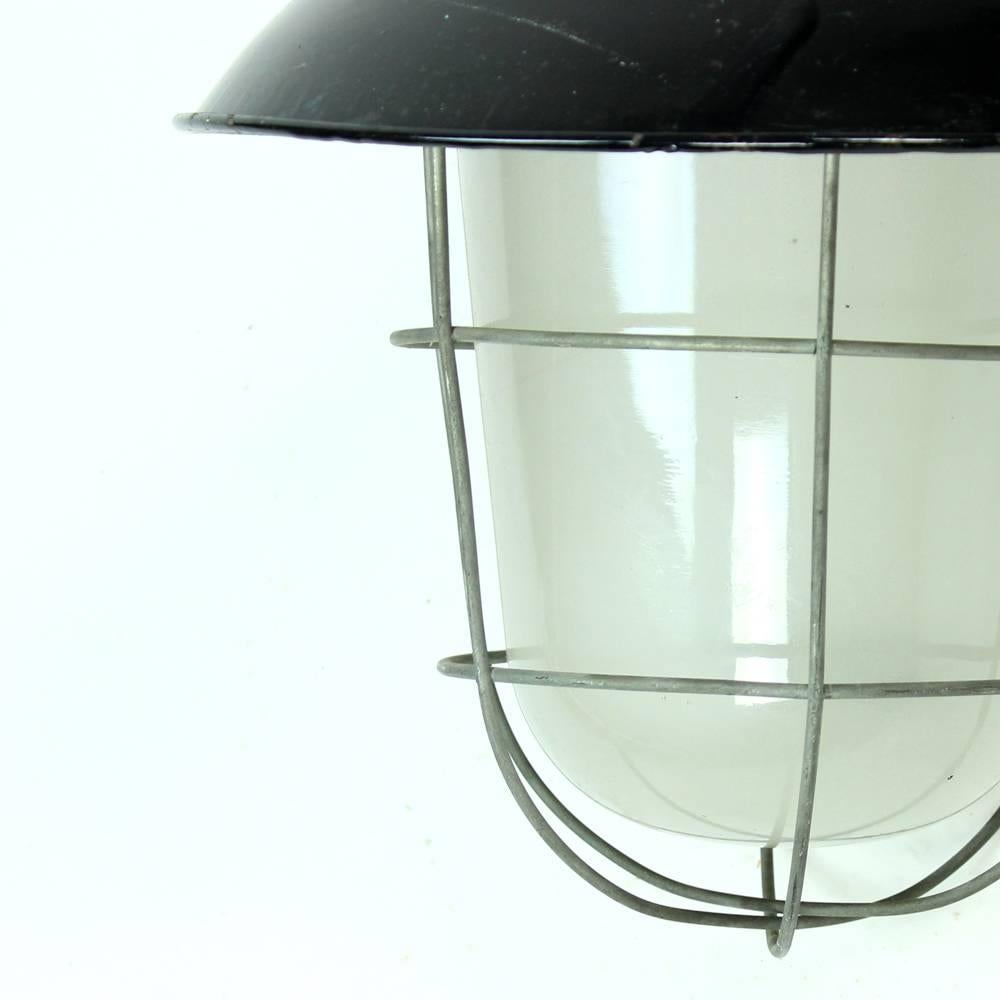 Industrial light with metal top in black emanel finish. Glass light covered and protected by metal cage. Used in industrial factories in the past, the light is an interesting item in many interiors today. Very good condition with some wear visible