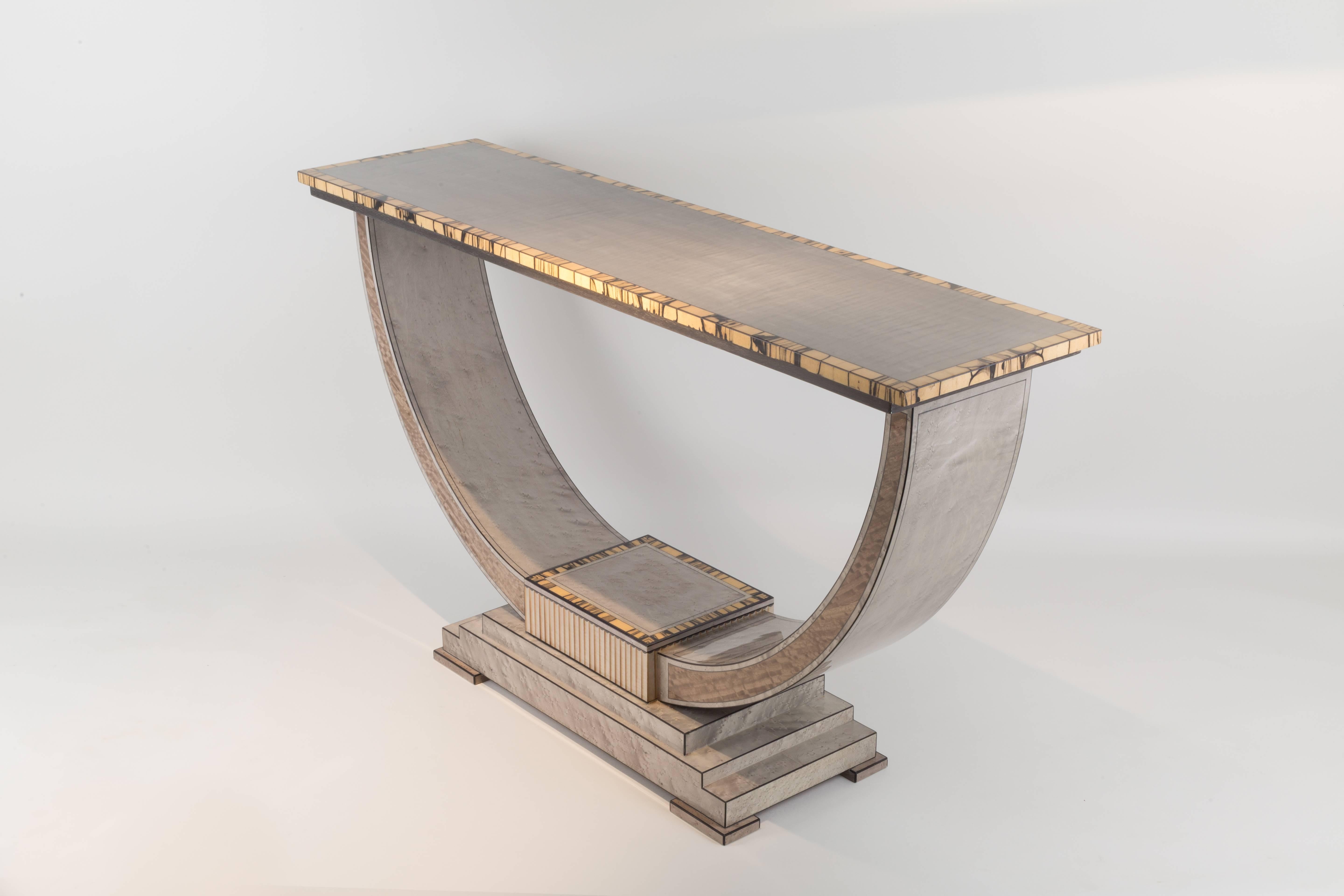 This exciting console table combines Italian sycamore and silver maple to stunning effect. Inlaid with black boxwood and crossbanded with white ebony.
Eucalyptus panels are set into the delicately tapered arch supports. The centre block is encased