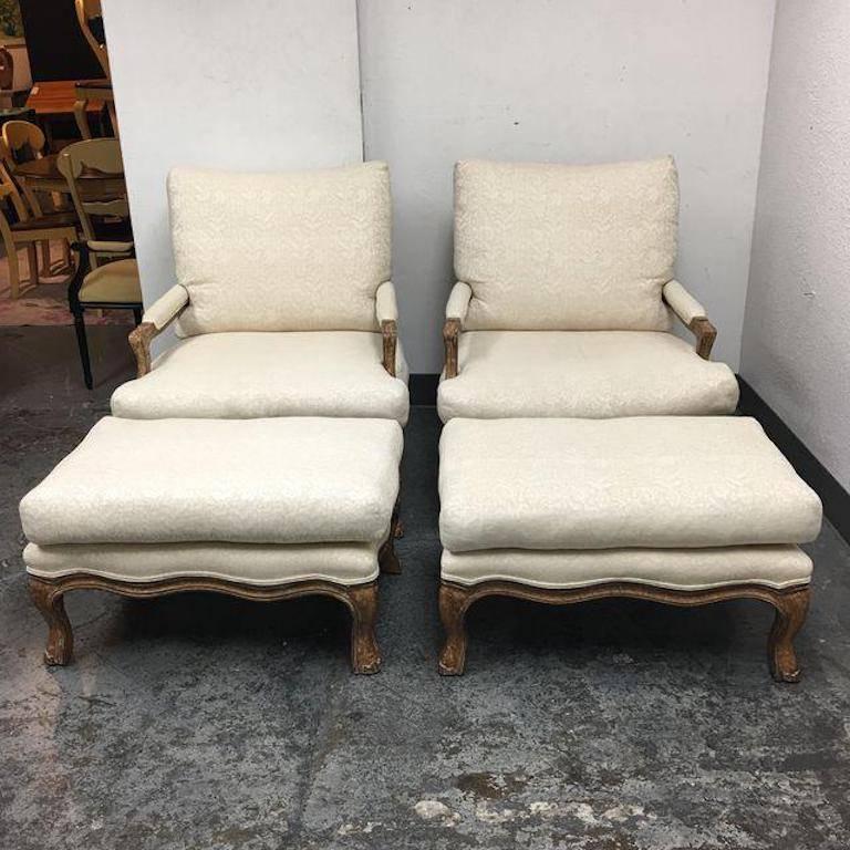 An oversized pair of Kreiss Collection Bergere armchairs that come with matching ottomans. The subtle carved wood frames are finished in a warm honey color that really sets off the almost demure Cream fabric with which they were upholstered. All of