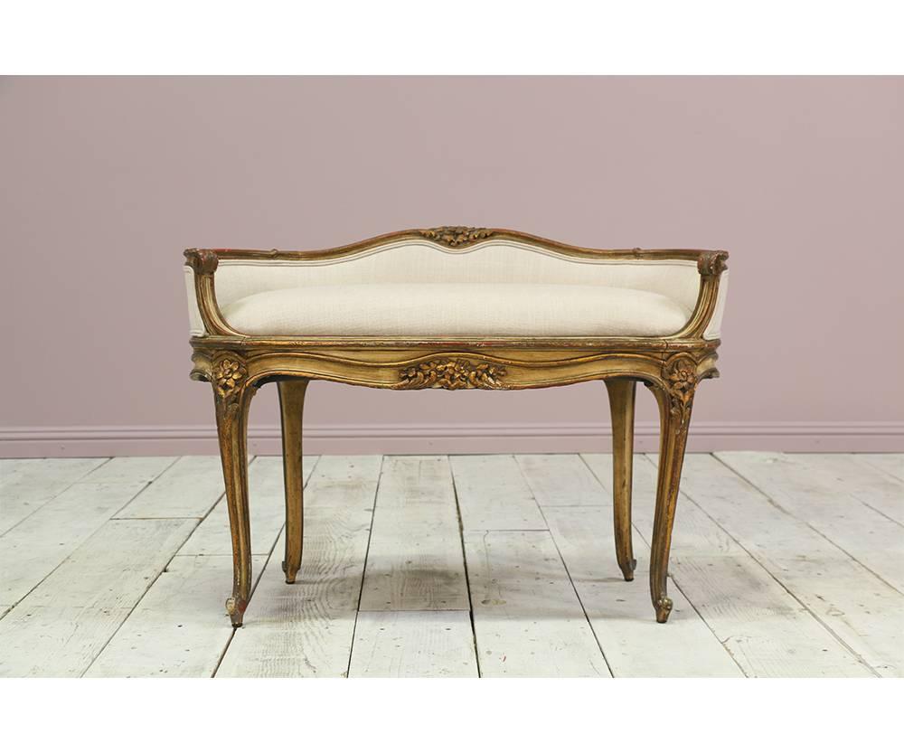  French 1920s Louis XV-style vanity bench featuring a wonderful naturally distressed painted and parcel-gilt finish with delicately carved floral motif decorations. The bench is sturdy and has been newly upholstered in a high quality linen fabric.