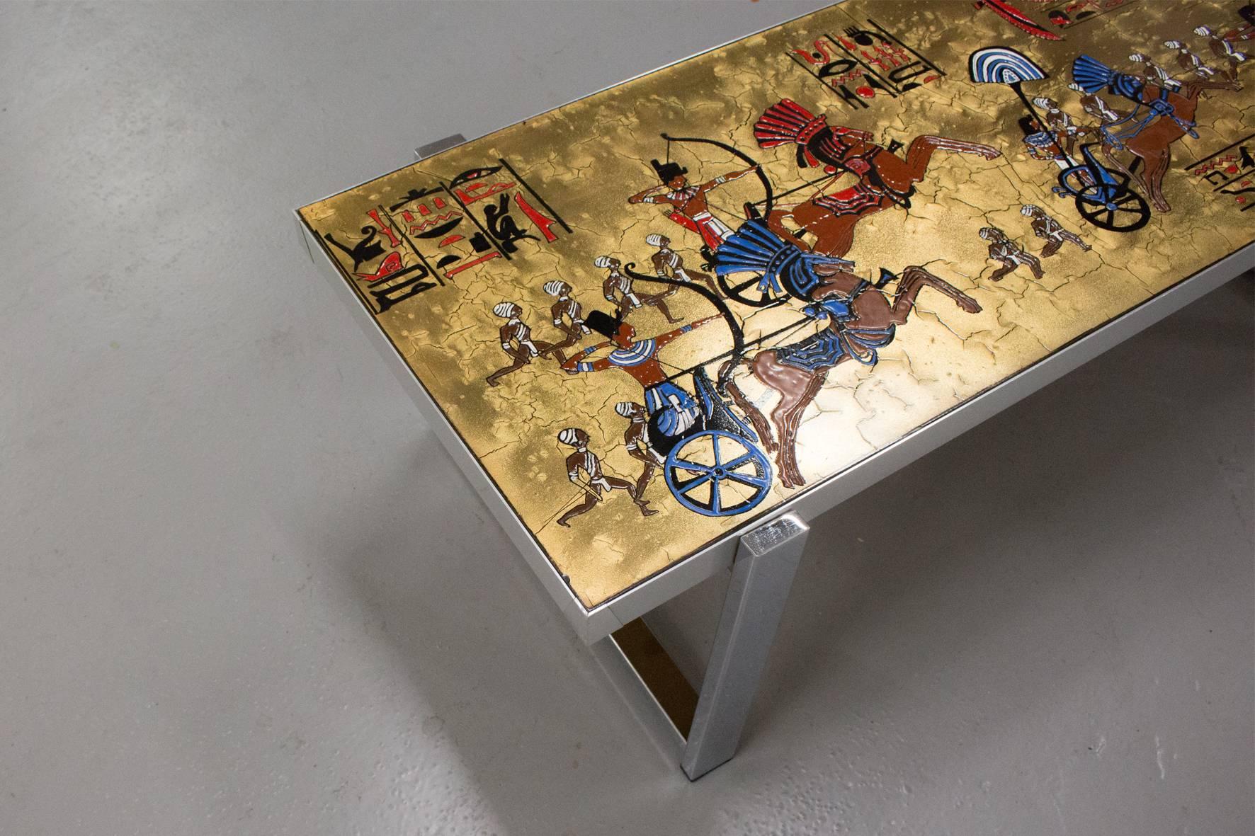 This signed Denisco Egyptian-themed coffee table was originally produced in Belgium in the 1960s or 1970s. The top is made of a golden hand-painted tile with a detailed Egyptian setting.

The sides and legs are made out of a glossy chrome, which