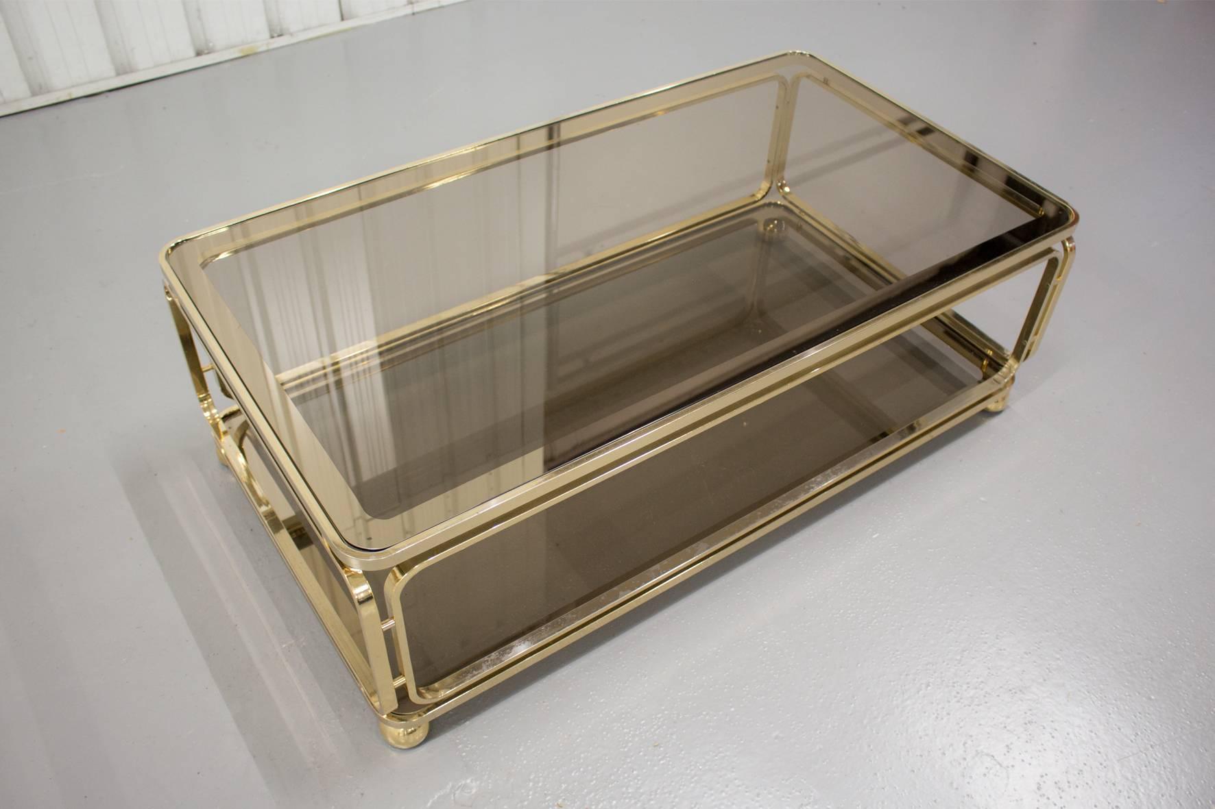 A beautiful Regency-style coffee table with two tiers made of smoked glass with a mirrored edge. Underneath the messing frame are 4 easily adjustable wheels to move the table in a comfortable manner. 

The frame has a little wear and a few minor