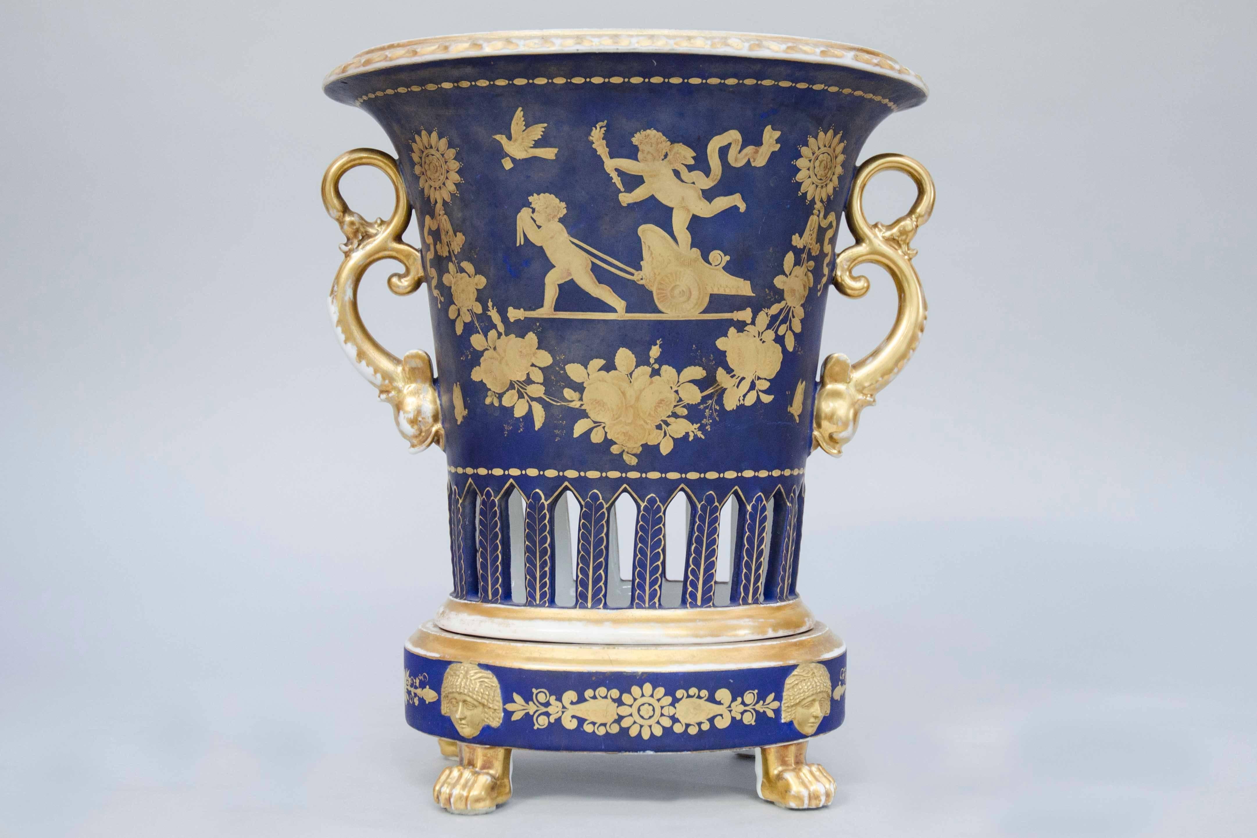 Pendulum clock in a porcelain urn with gilded neoclassical decoration, like horns of plenty and angles riding a chariot to deliver the torch of passion and a love letter. Deep blue ground. Dolphins handles. Paw feet. Original movement and enamel