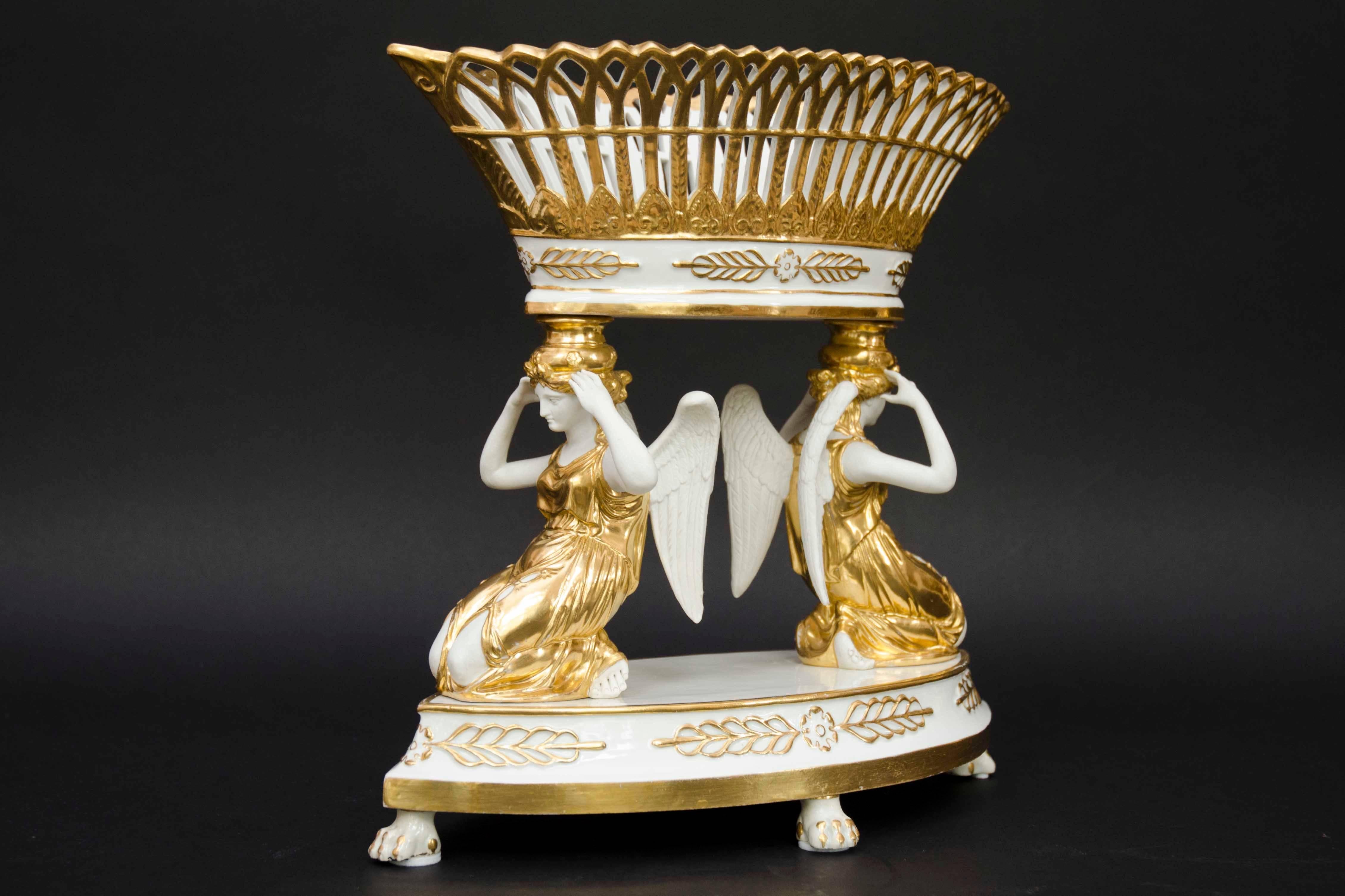 Large porcelain open worked fruit basket in the Empire style held by a couple of winged women. Oval shape, women kneeling down with their arms holding the basket on their heads. The figures are in bisque. Highlights in gold on the porcelain and the