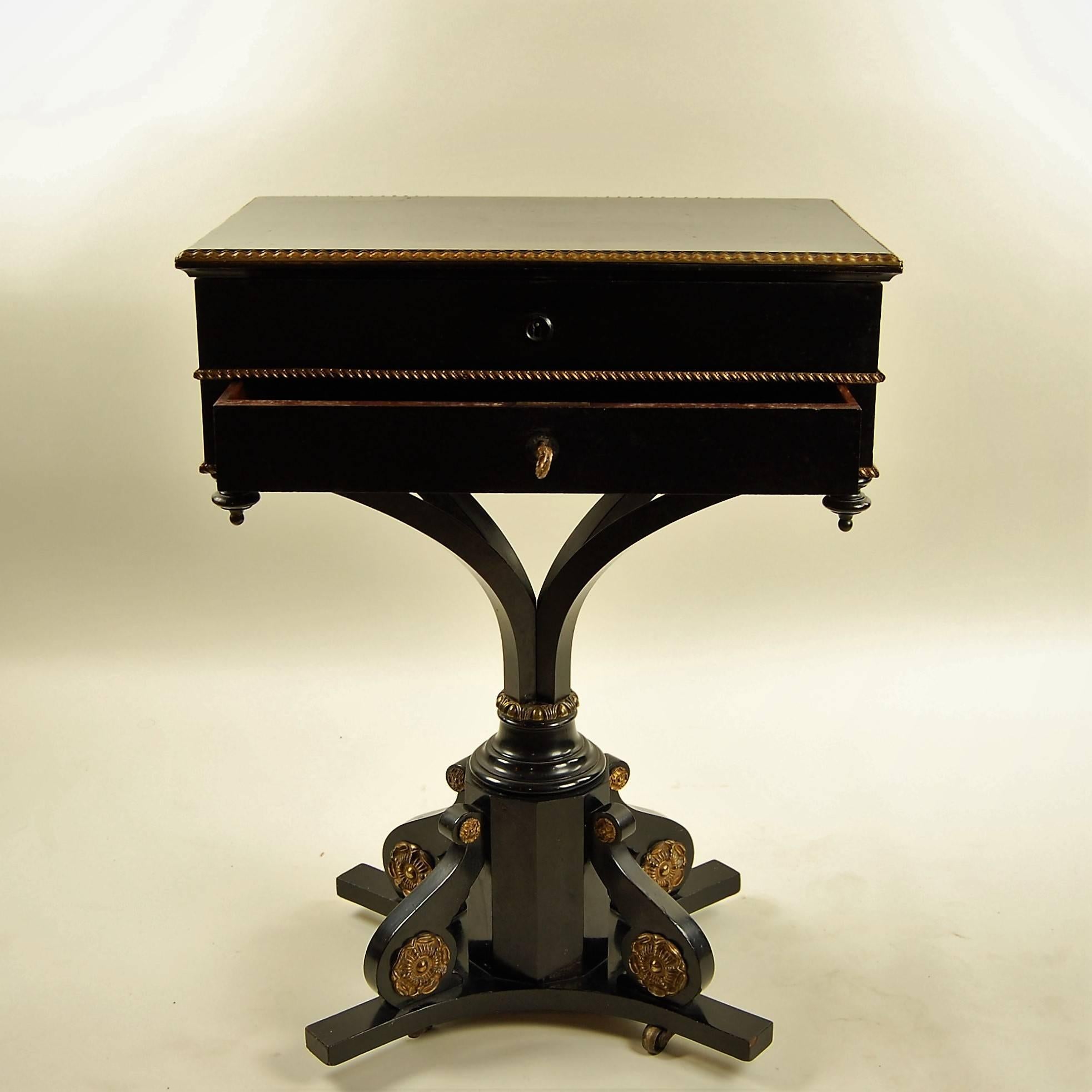A useful accessory for a 19th-century parlor or drawing room, this charming little table features a rich ebony finish with gilded accents. Rosette detail add interest to the bottom center pedestal. Two locking drawers (key included) provide access