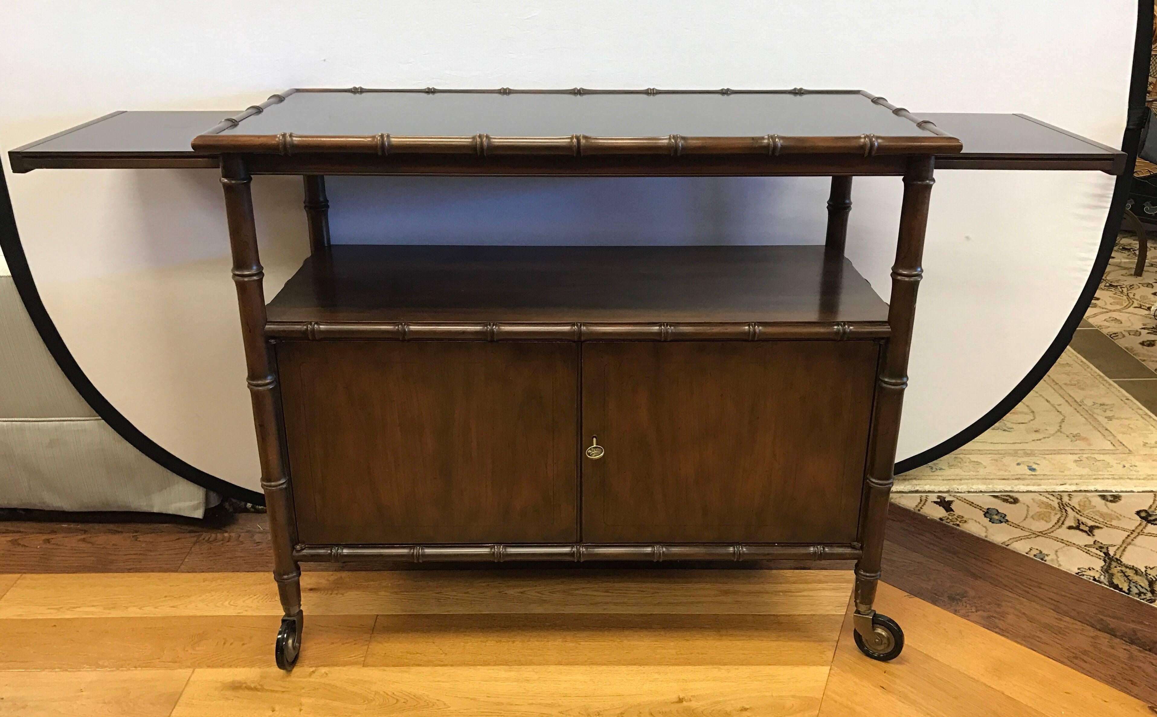 Midcentury chinoiserie faux bamboo rolling bar cart with pull-out sides that expand to 64 inches. Bottom has two doors with working lock and key. Top is black laminate. Baker medallion inside left drawer.