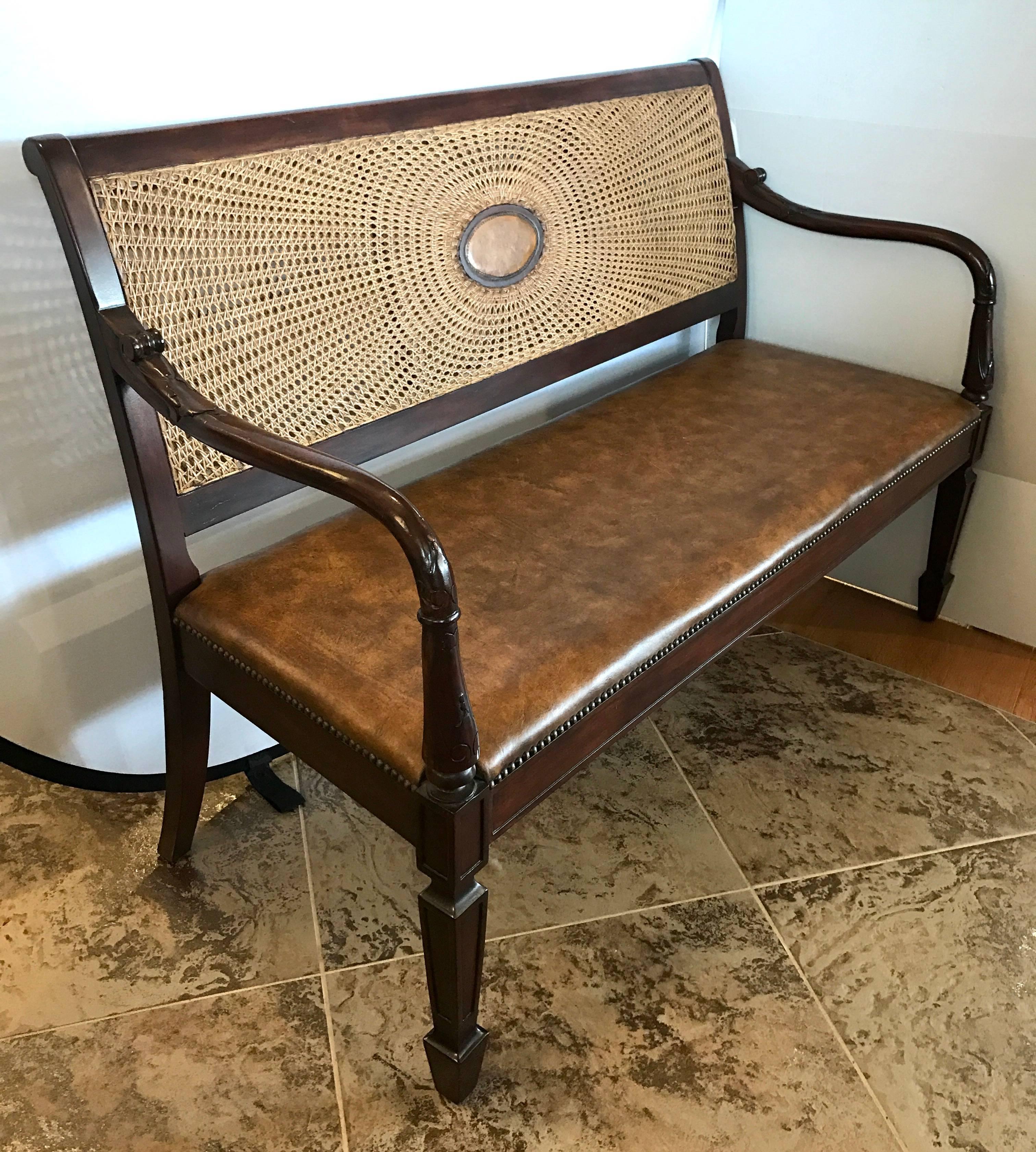 Unique British Colonial mahogany bench with carved arms and a double layered caned back featuring unusual medallion. Seat is upholstered in a brown leather and finished with brass nailheads.