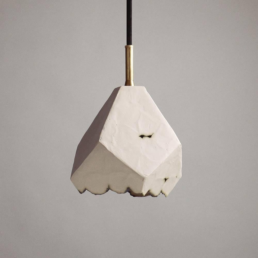 This sculptural pendant lamp features a clean geometric shape and a unique textural matte finish. The shade is handcrafted from slabs of unglazed white porcelain with highly individual black oxide burnout detailing at the edges. An unfinished brass
