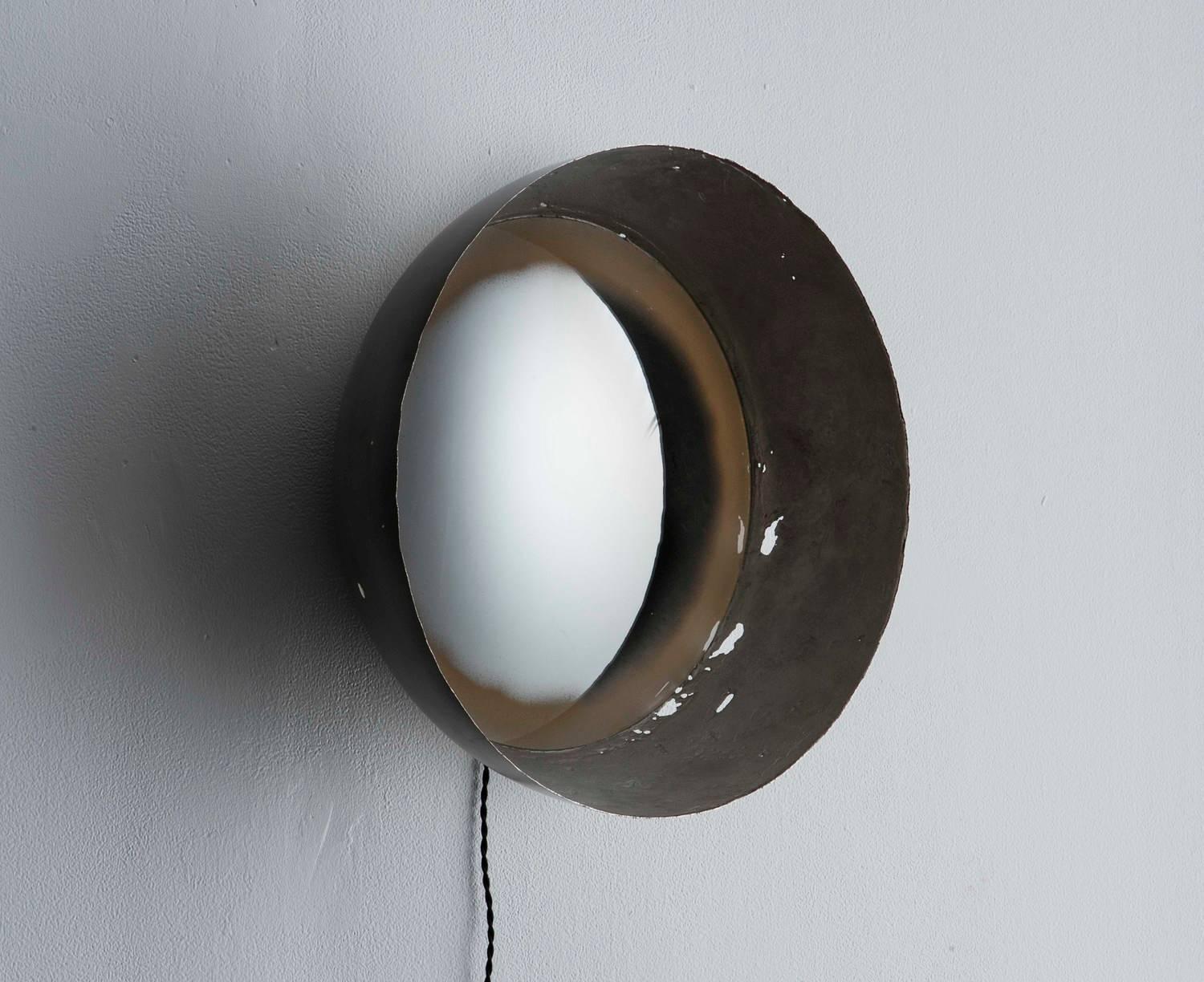 This curved light fixture creates a stunning lighting effect using various curved surfaces. The body of the light is a formed metal hemisphere with an inset convex mirror. The edge of the mirror is backlit through an area with a hand applied