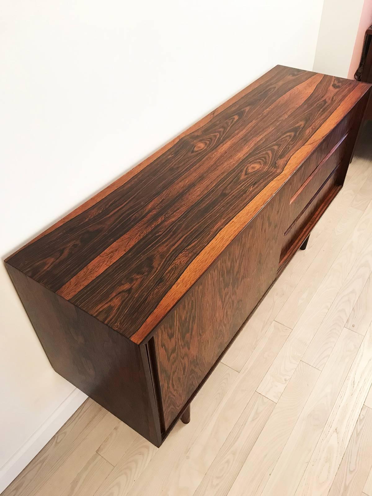 Super cute and petite rosewood server. Made in Denmark by Steens. Three drawers and one sliding cabinet with shelve inside. Pretty rosewood turned peg legs. The rosewood has a very pretty grain. Excellent condition with one chunk out of the veneer