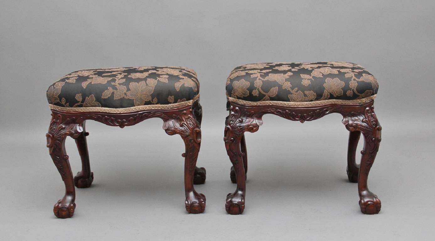 A decorative pair of mid-20th century mahogany stools, each of serpentine outline, upholstered in a dark colored fabric with a floral pattern, supported on foliate carved cabriole legs terminating in claw and ball feet, circa 1950.