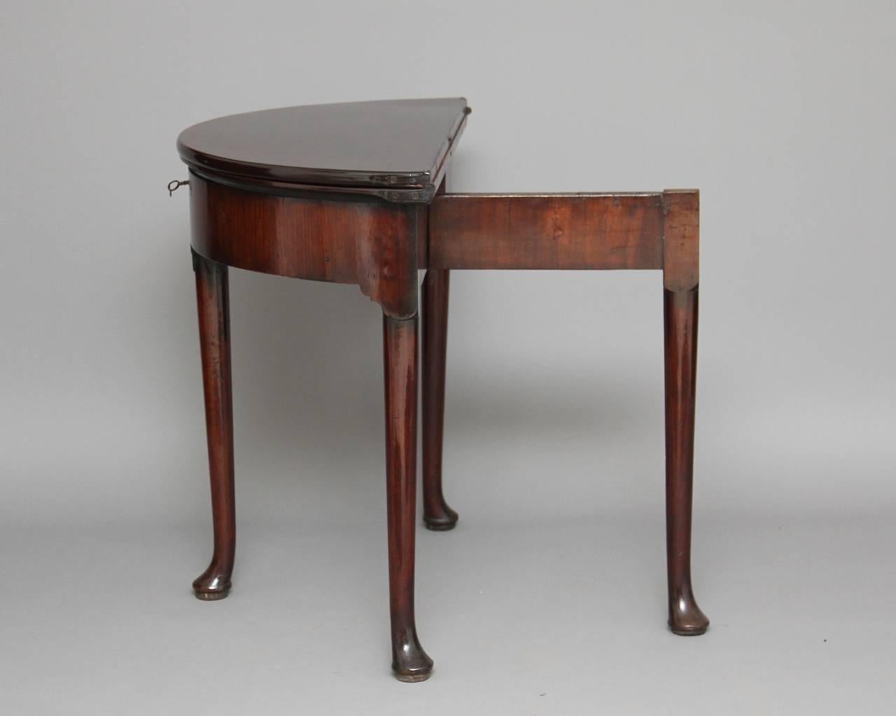 18th century mahogany demilune card/ games table, top folding over to reveal a playing surface lined with a red baize with guinea wells, then folding over the other way to reveal a compartment, standing on slender turned legs with pad feet, circa